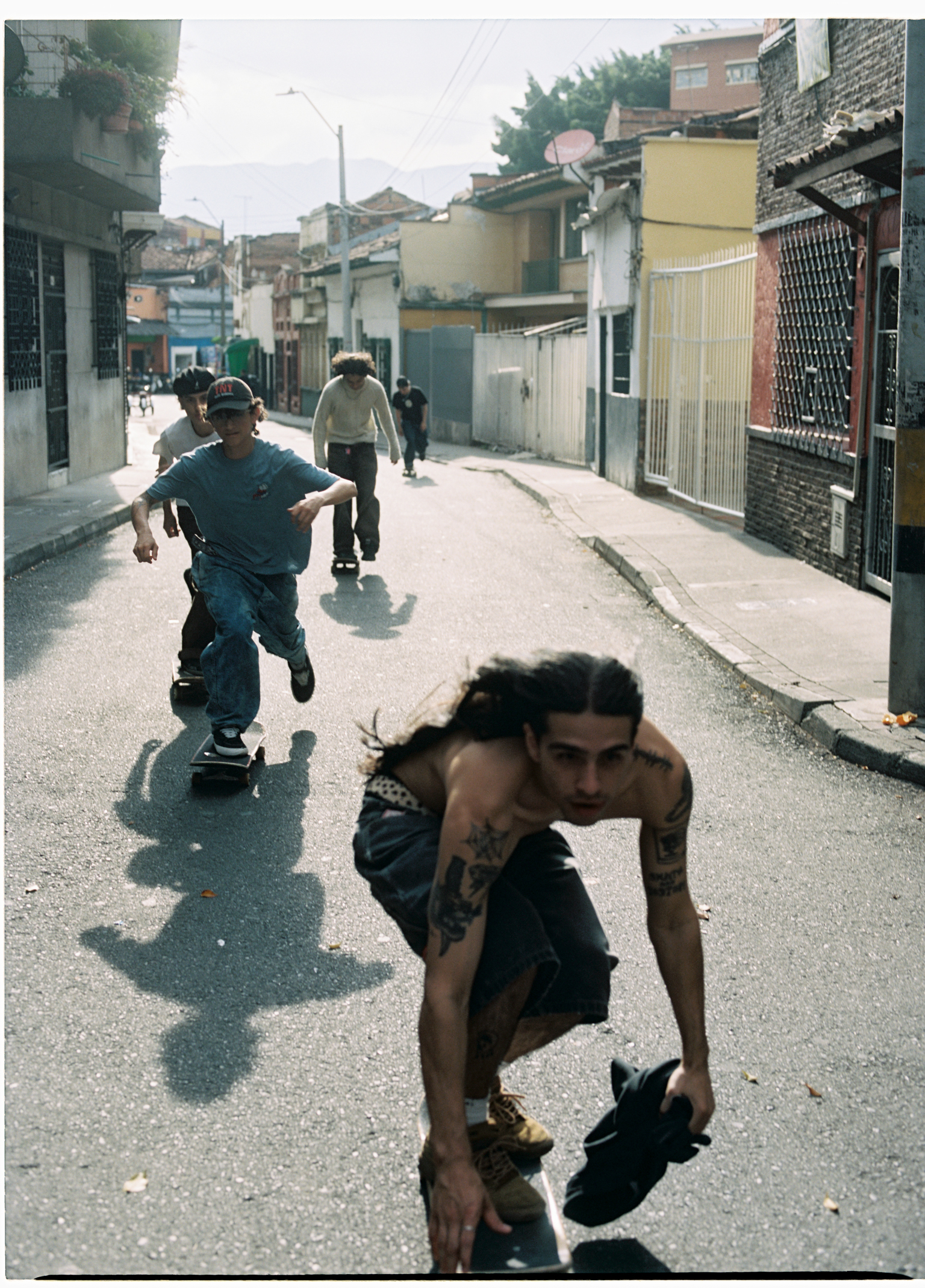 Young people skateboarding on a paved street lined with low-rise buildings and shops in bright daylight.