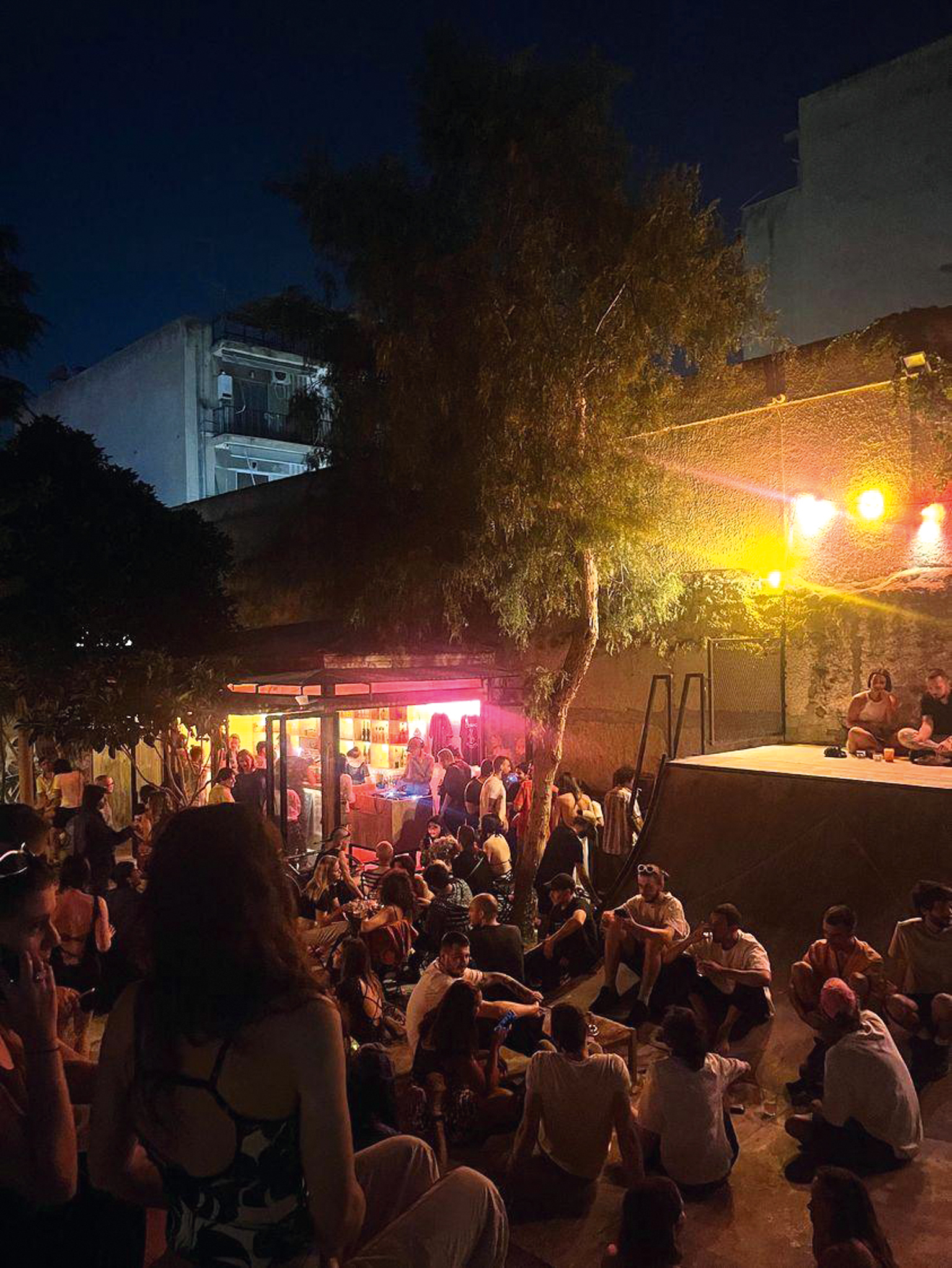 Crowded evening street scene with people gathered around illuminated food stall. Pink and yellow lights, dark blue sky, trees and buildings visible.