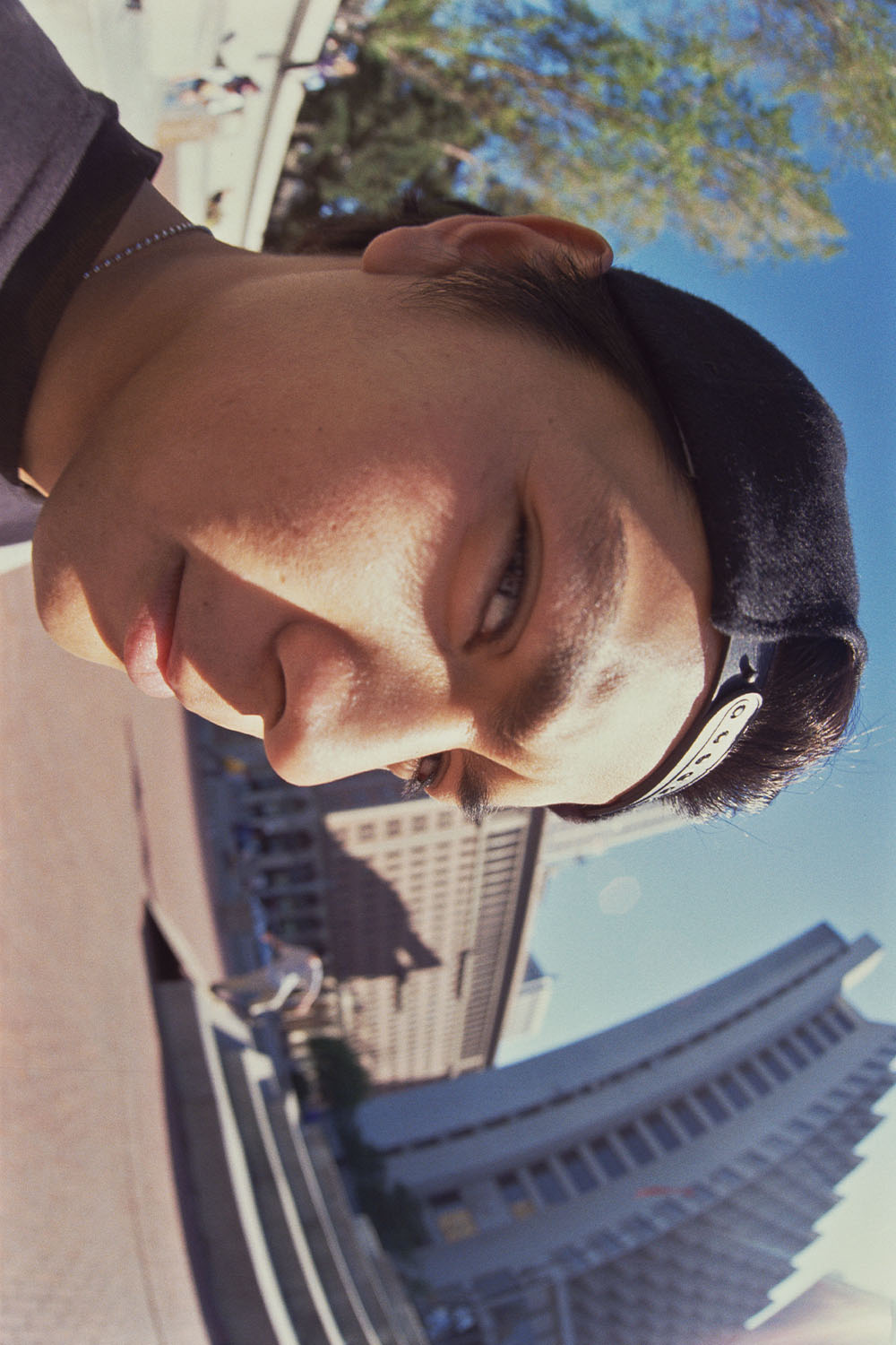 Man with dark hair and beard smiling, holding phone for selfie. Urban buildings and blue sky with trees visible in background.