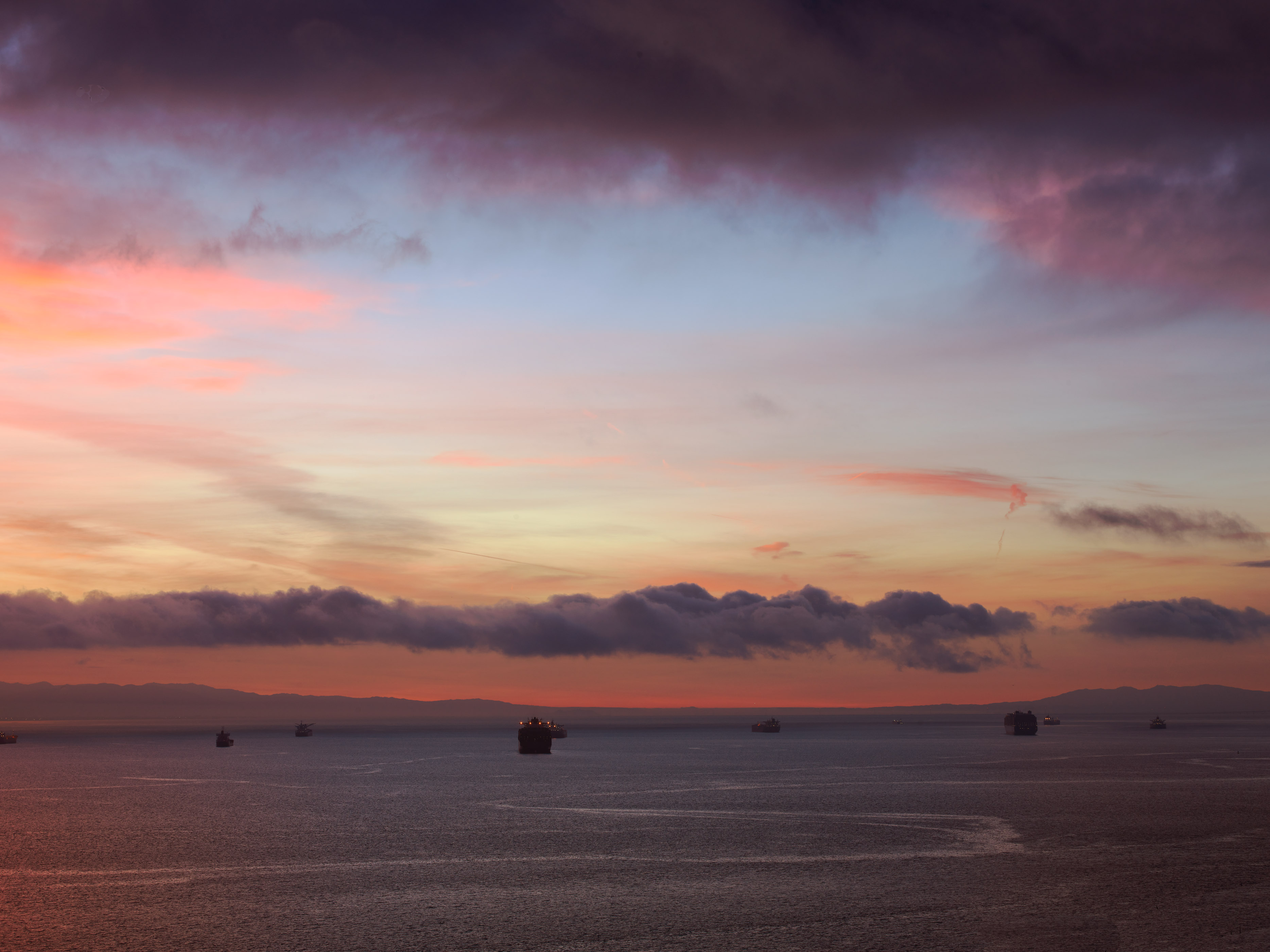 Dark sea with several boats scattered across horizon. Sky displays purple clouds above, orange and pink hues in middle, pale yellow below.