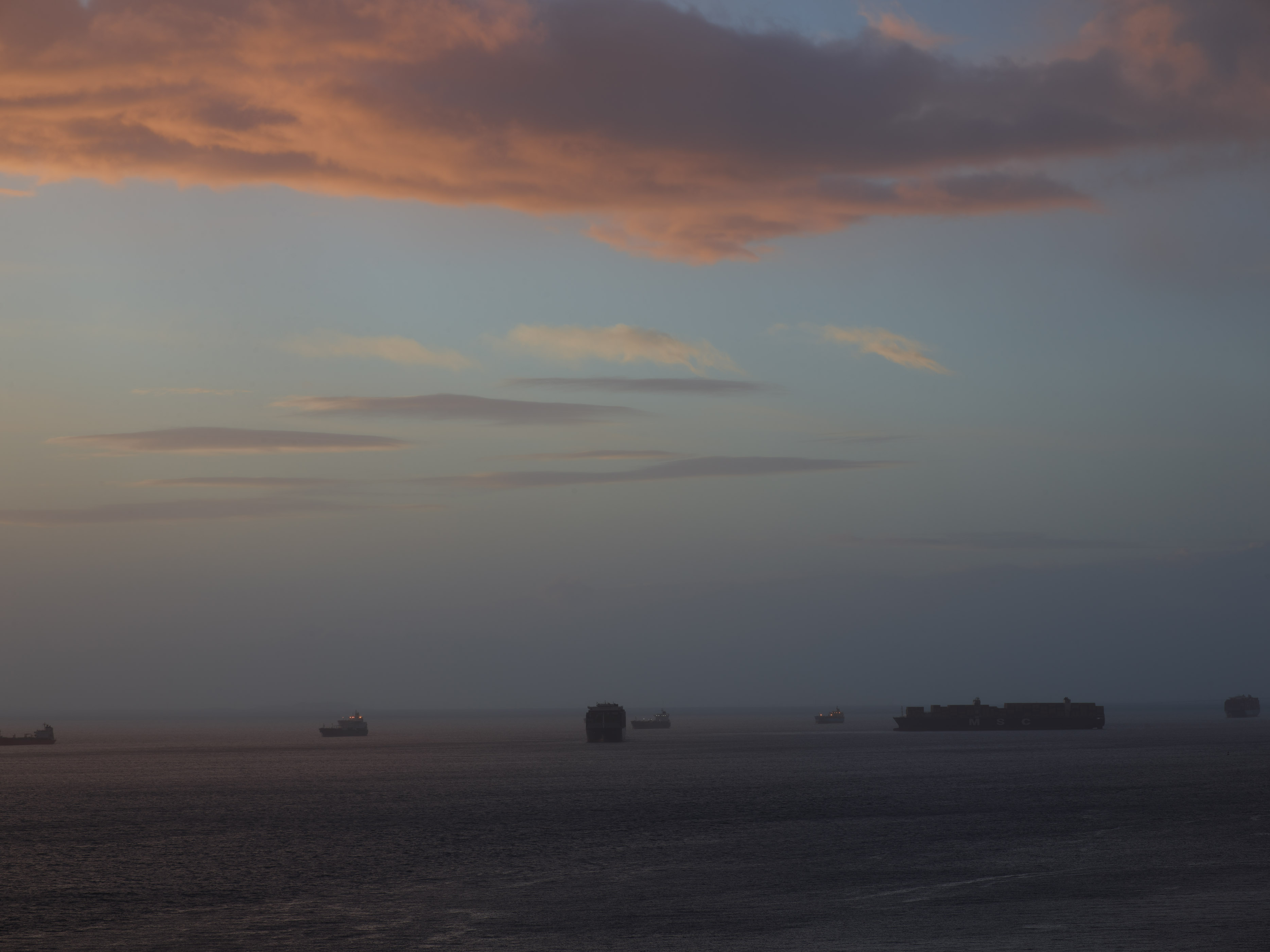 Ocean view with ships on horizon under cloudy sky with orange and pink sunset colours reflecting on water.