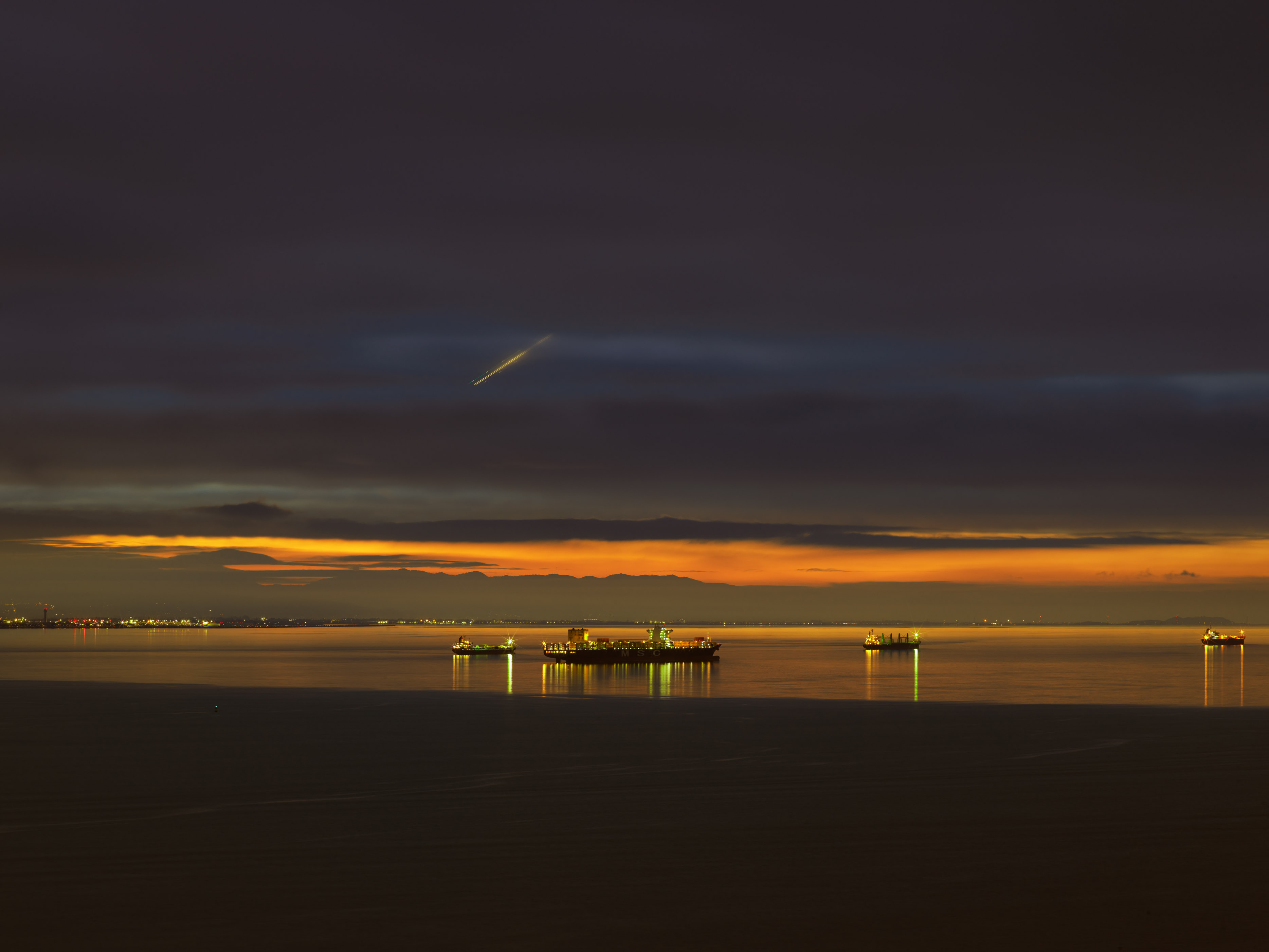 Dark seascape with orange sunset strip along horizon. Purple-grey sky above, black water below with scattered boat lights reflecting on surface.