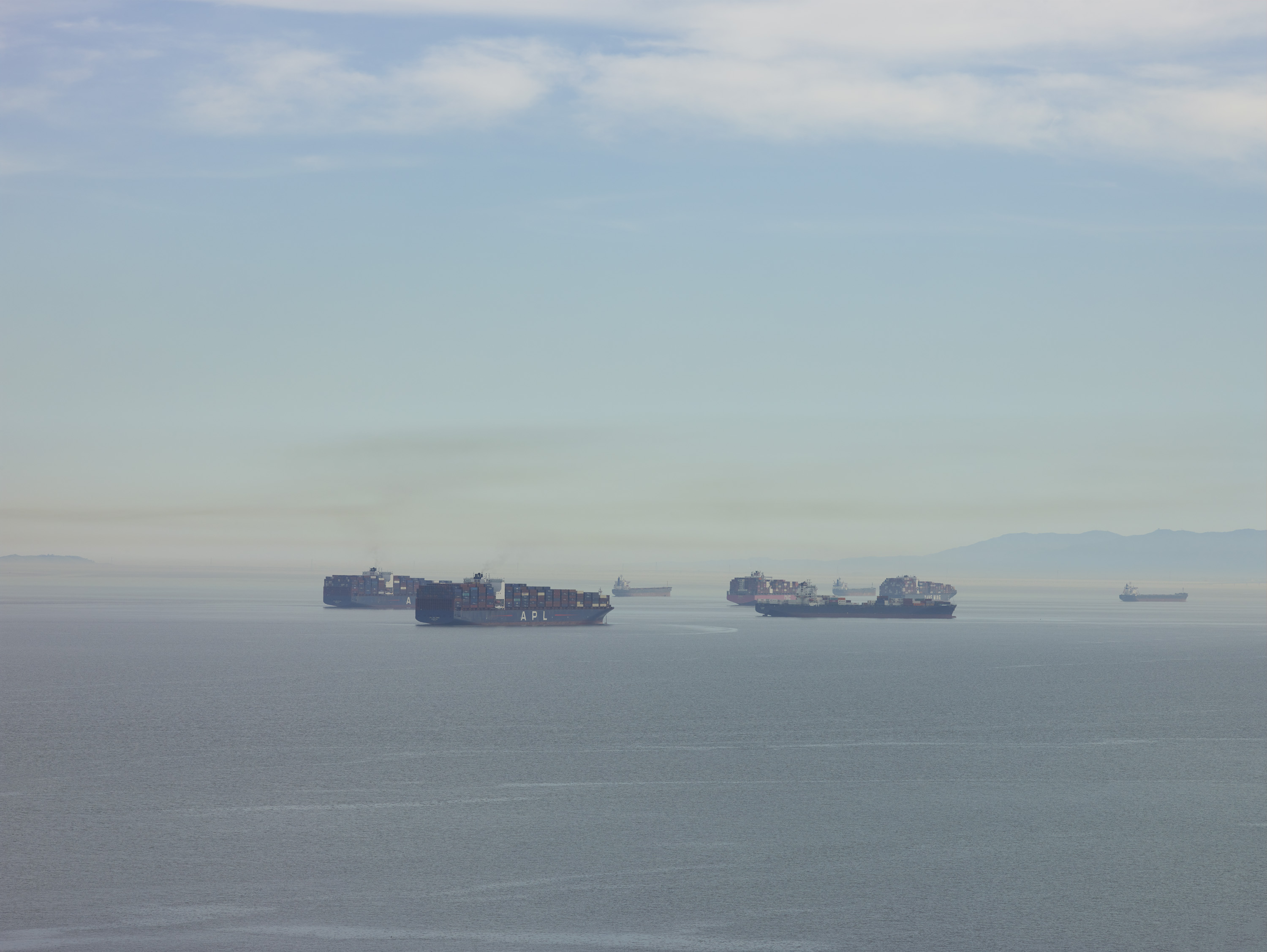 Several dark ships silhouetted against grey water under pale blue sky with white and grey clouds.