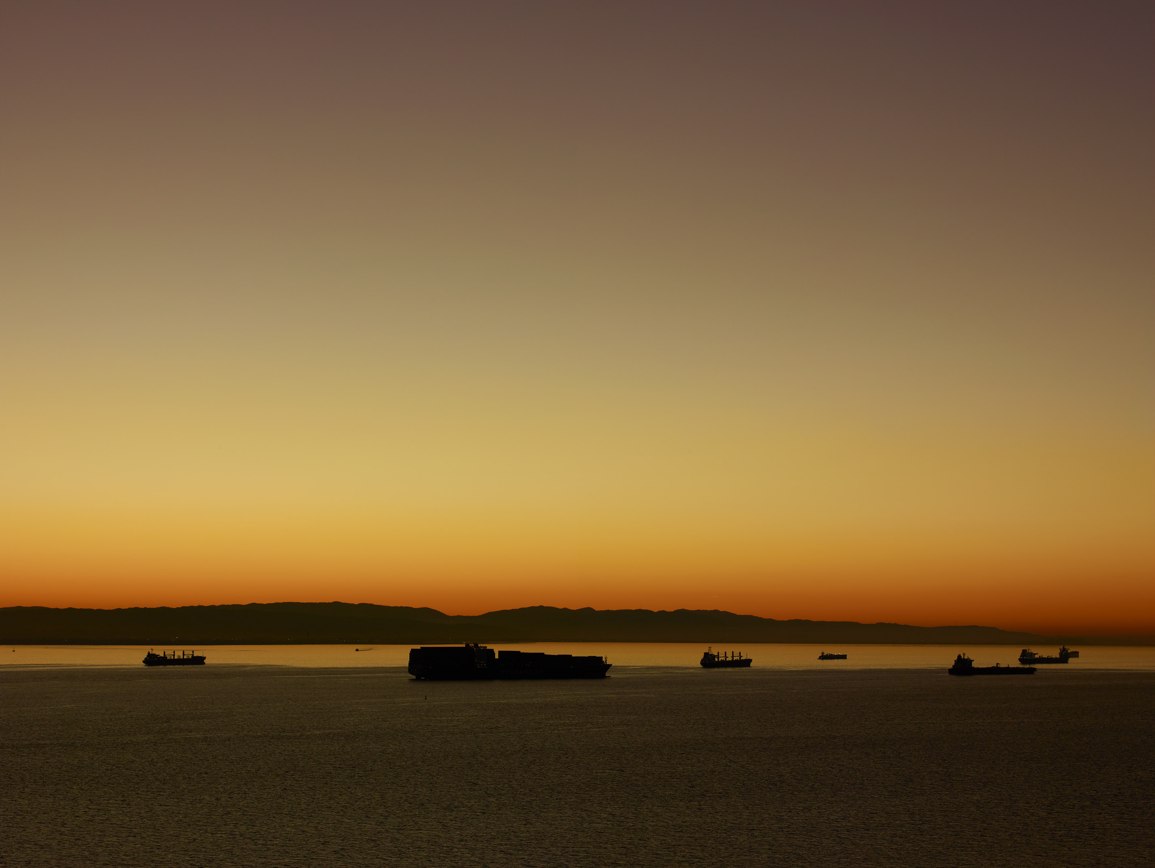 Ships silhouetted on calm water against orange and yellow gradient sky transitioning to purple clouds above distant shoreline.