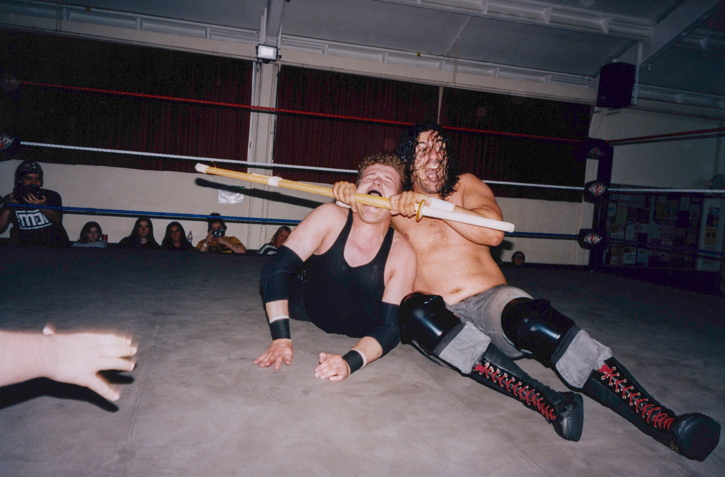 Two wrestlers grappling on mat in boxing ring, one applying chokehold from behind whilst spectators watch from ringside seating.