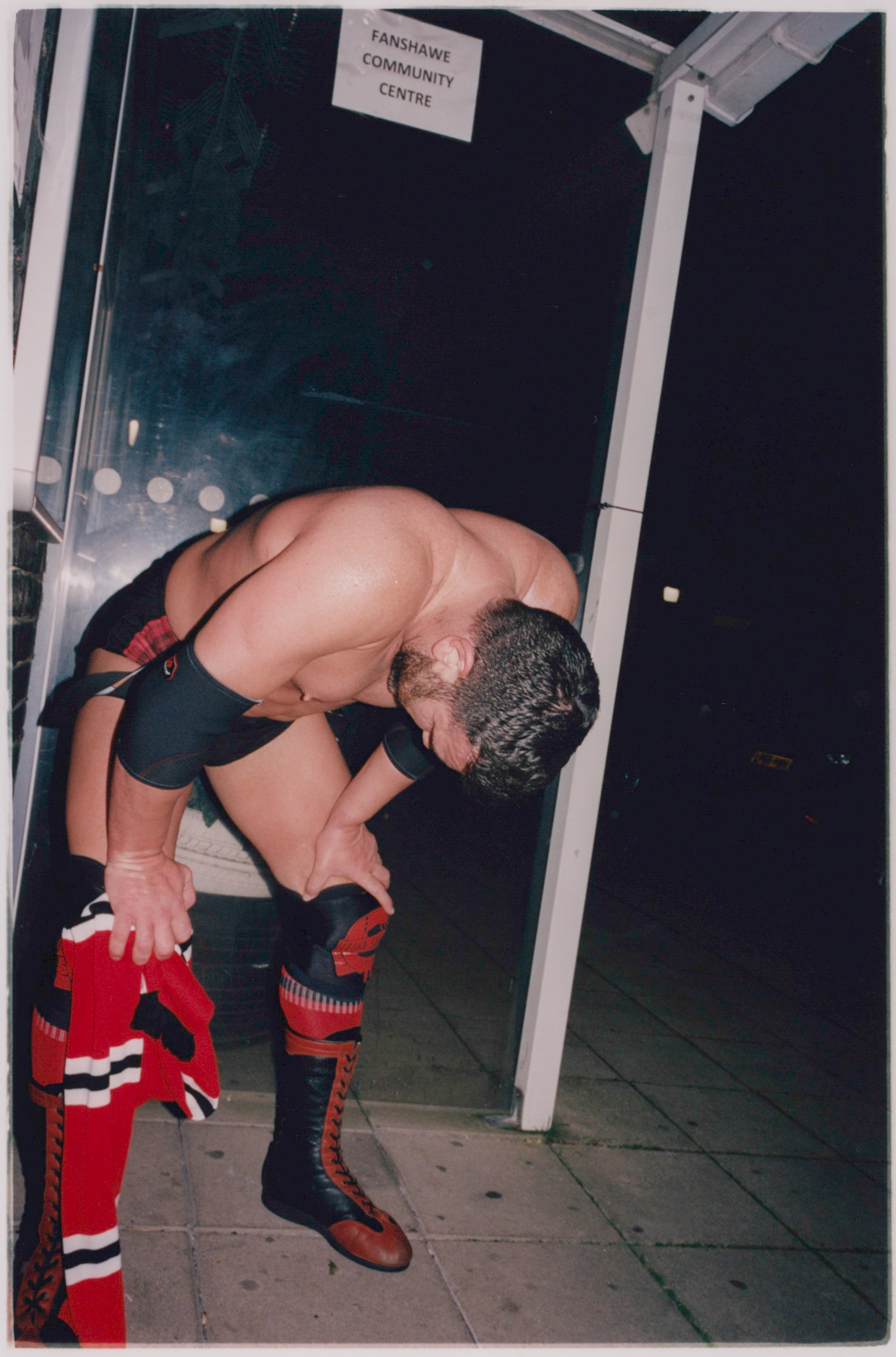 Shirtless wrestler in black trunks and red boots bending forward in doorway of community centre building at night.