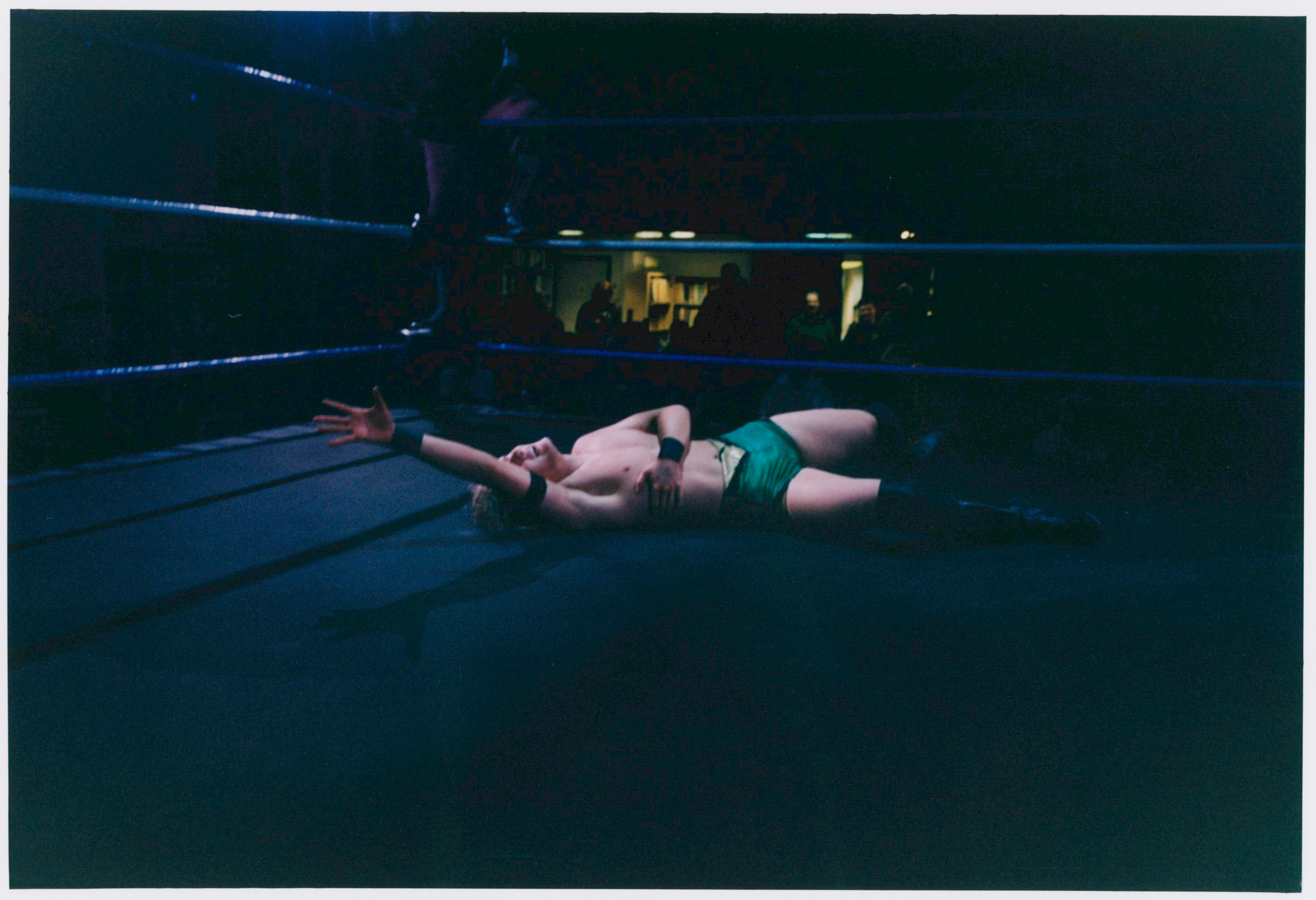 Wrestler in white trunks lying on dark blue wrestling ring mat, with ropes visible and yellow lighting in background.