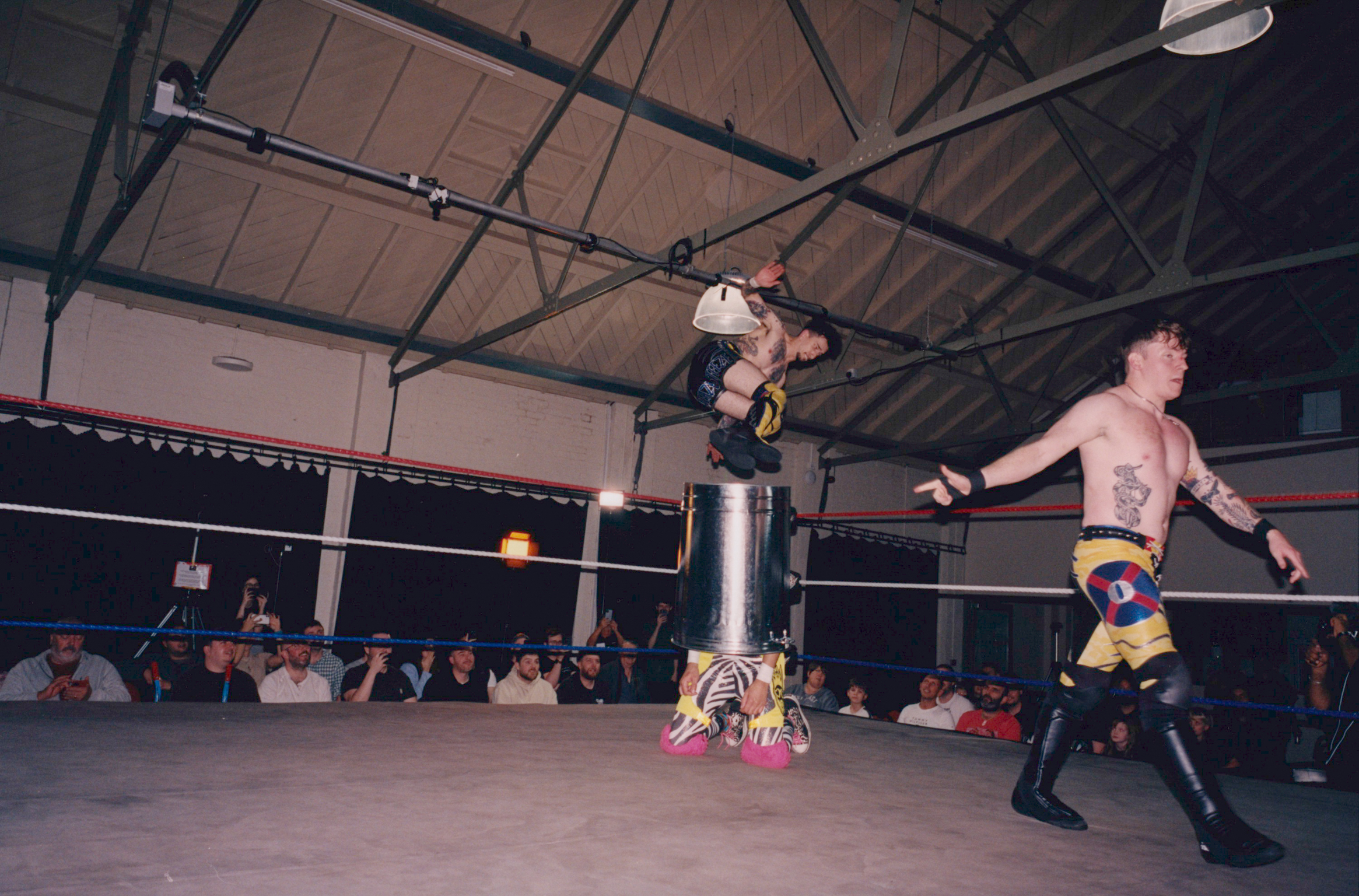 Professional wrestling match in indoor venue with one wrestler mid-air performing aerial move whilst opponent stands in ring corner.