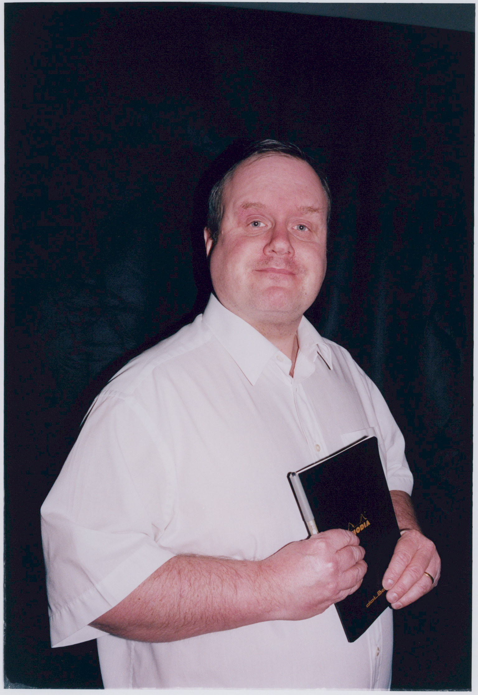 Man in white shirt holding black book, standing against dark blue-green background.