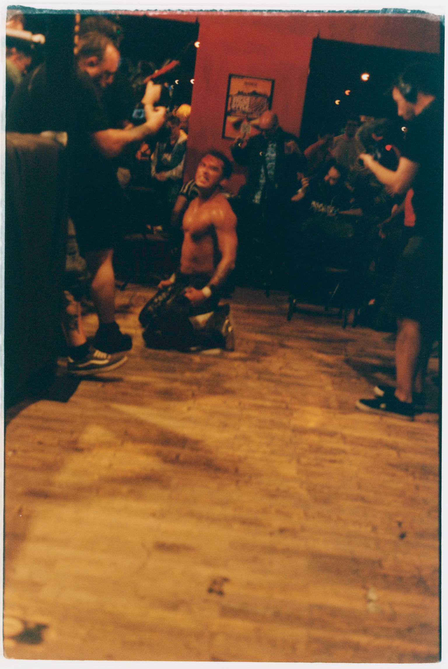 Band performing in dimly lit room with wooden floor. Musicians hold guitars, one person kneels centre stage. Warm orange lighting.