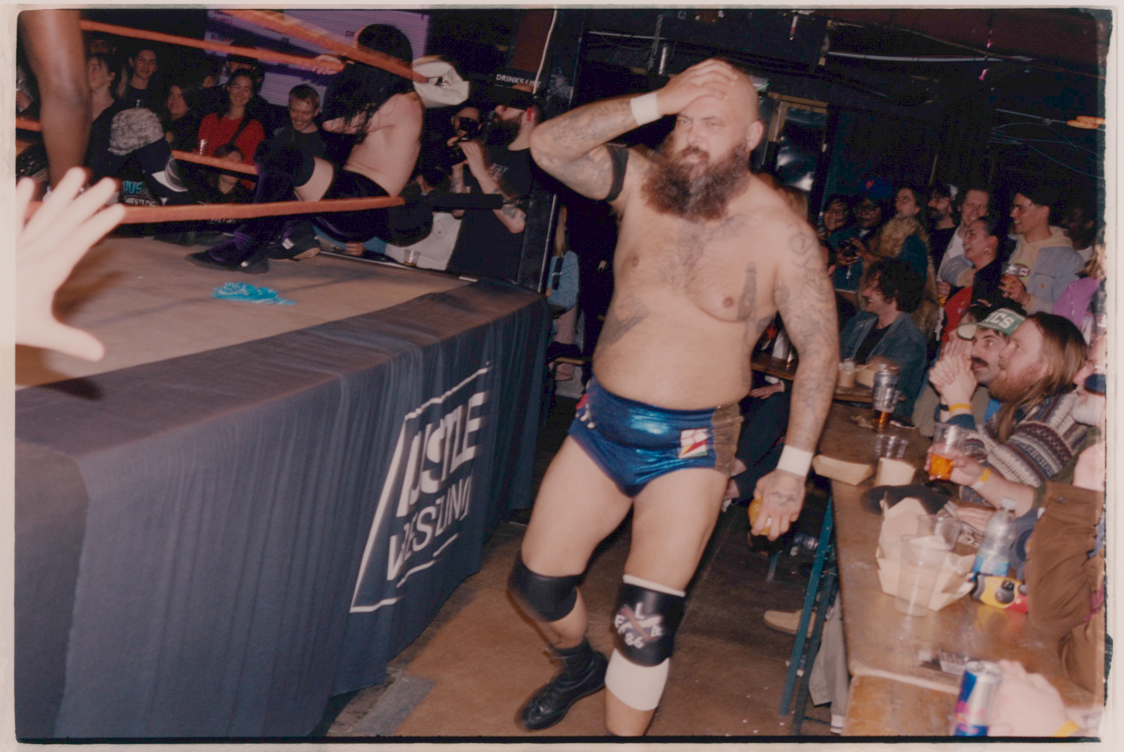 Shirtless wrestler with beard in blue trunks and black boots stands on wrestling ring apron, crowd watching from ringside seats.
