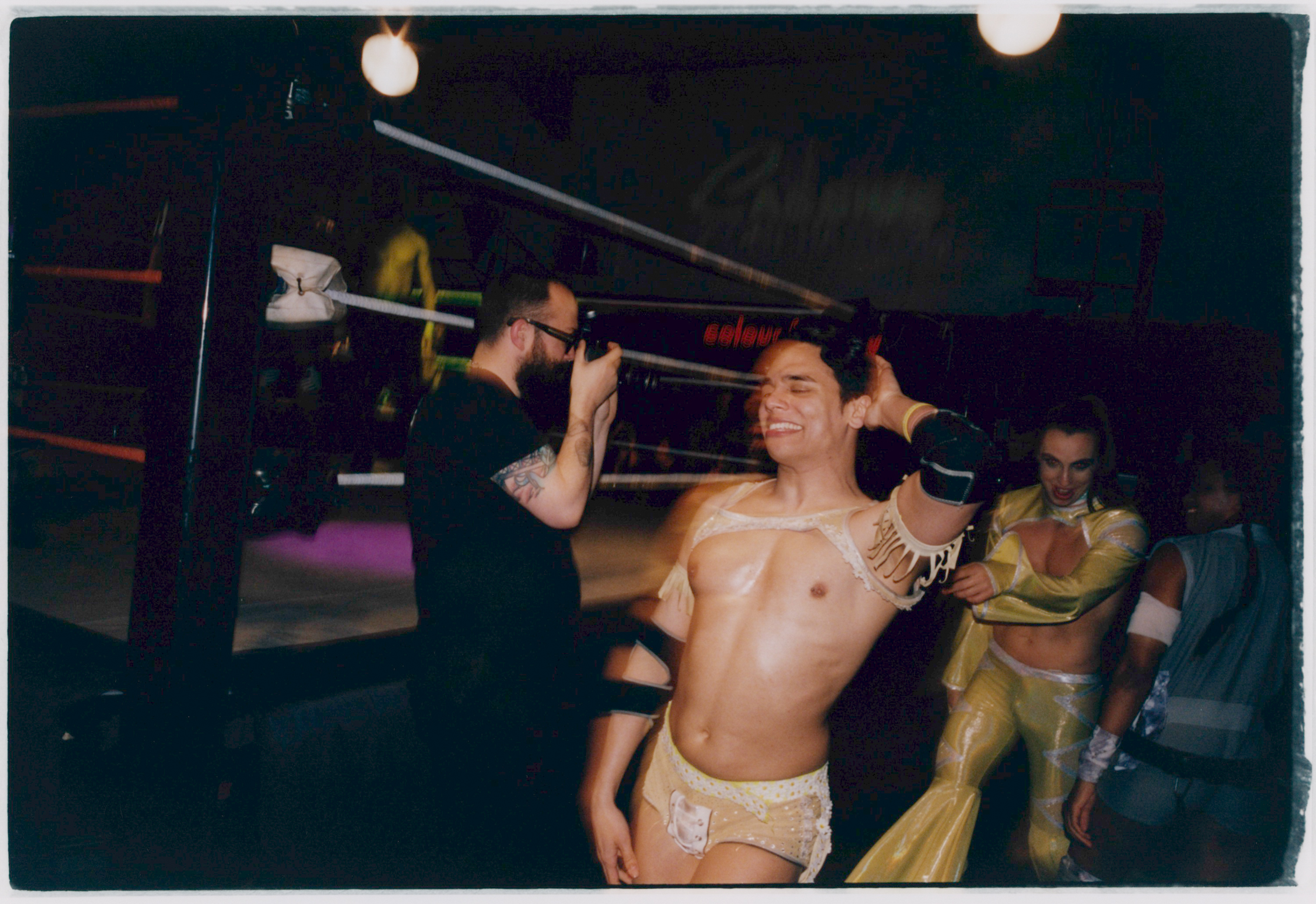 Shirtless man in white briefs flexing biceps in boxing ring, surrounded by people in dark indoor venue with overhead lighting.