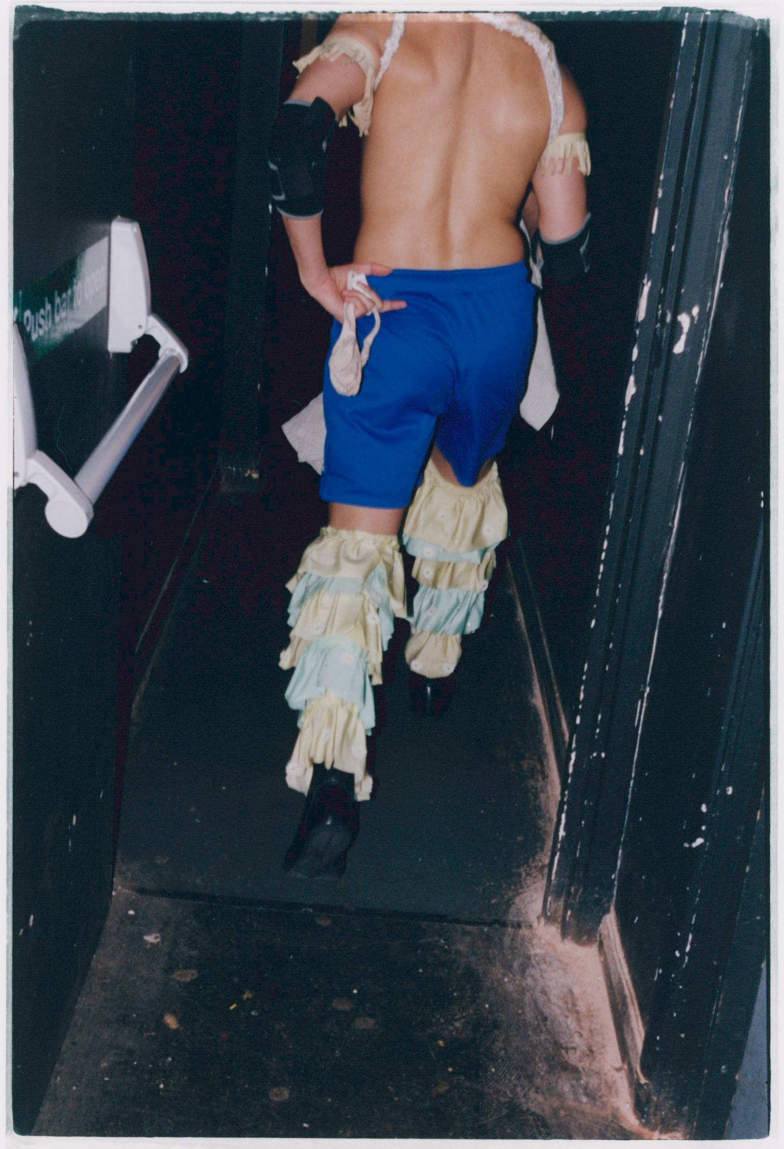 Shirtless person in blue shorts and fringed boots walking down dark corridor with handrail, taken from behind.
