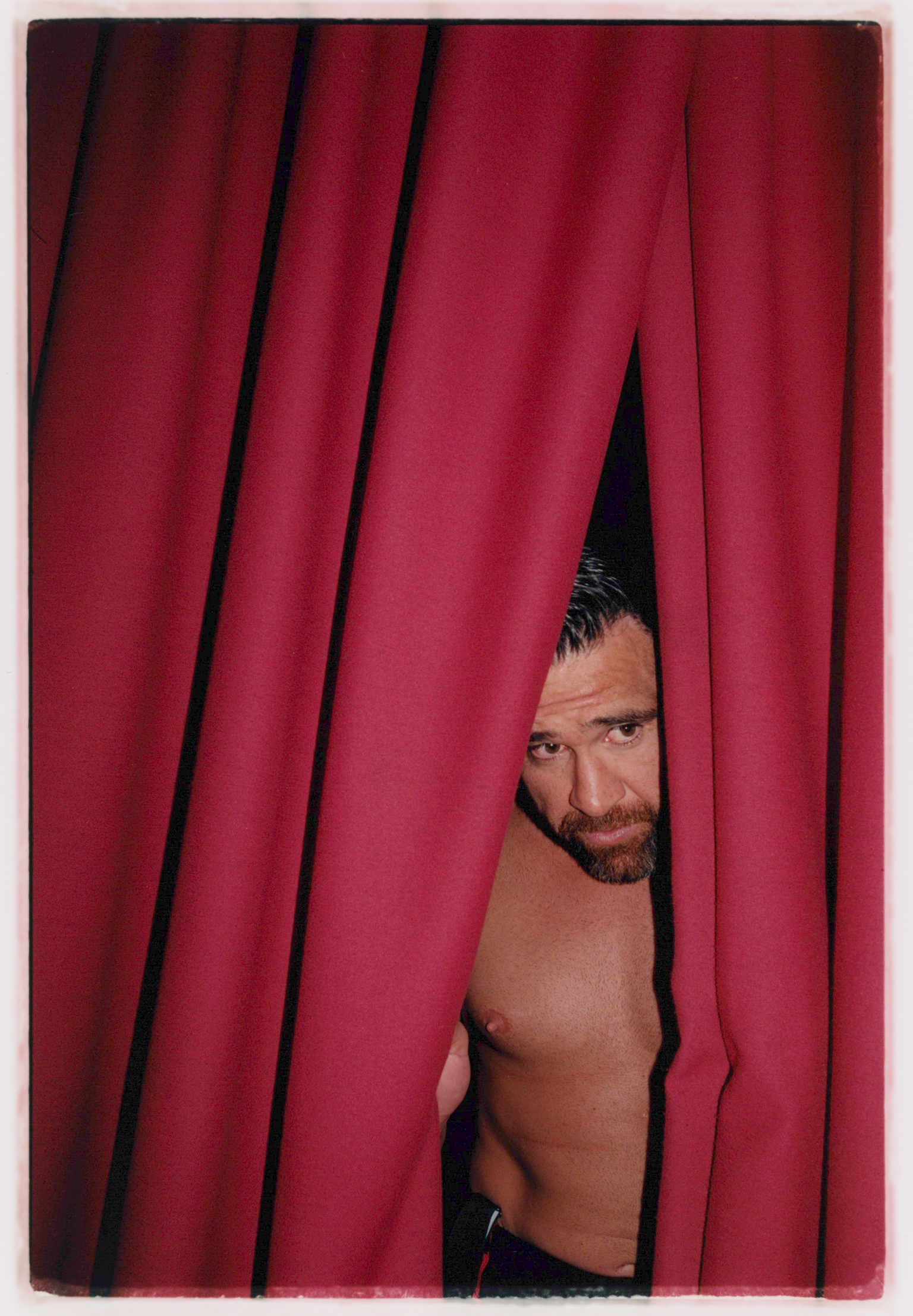Shirtless man with dark hair and beard peering through gap in deep red curtains, showing partial torso and face.