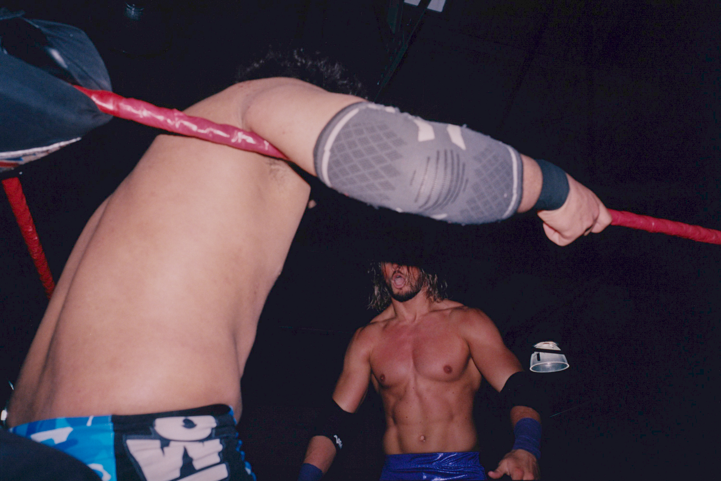 Two wrestlers in a ring, one shirtless in blue trunks, another leaning on red ropes wearing white knee pads and dark trunks.