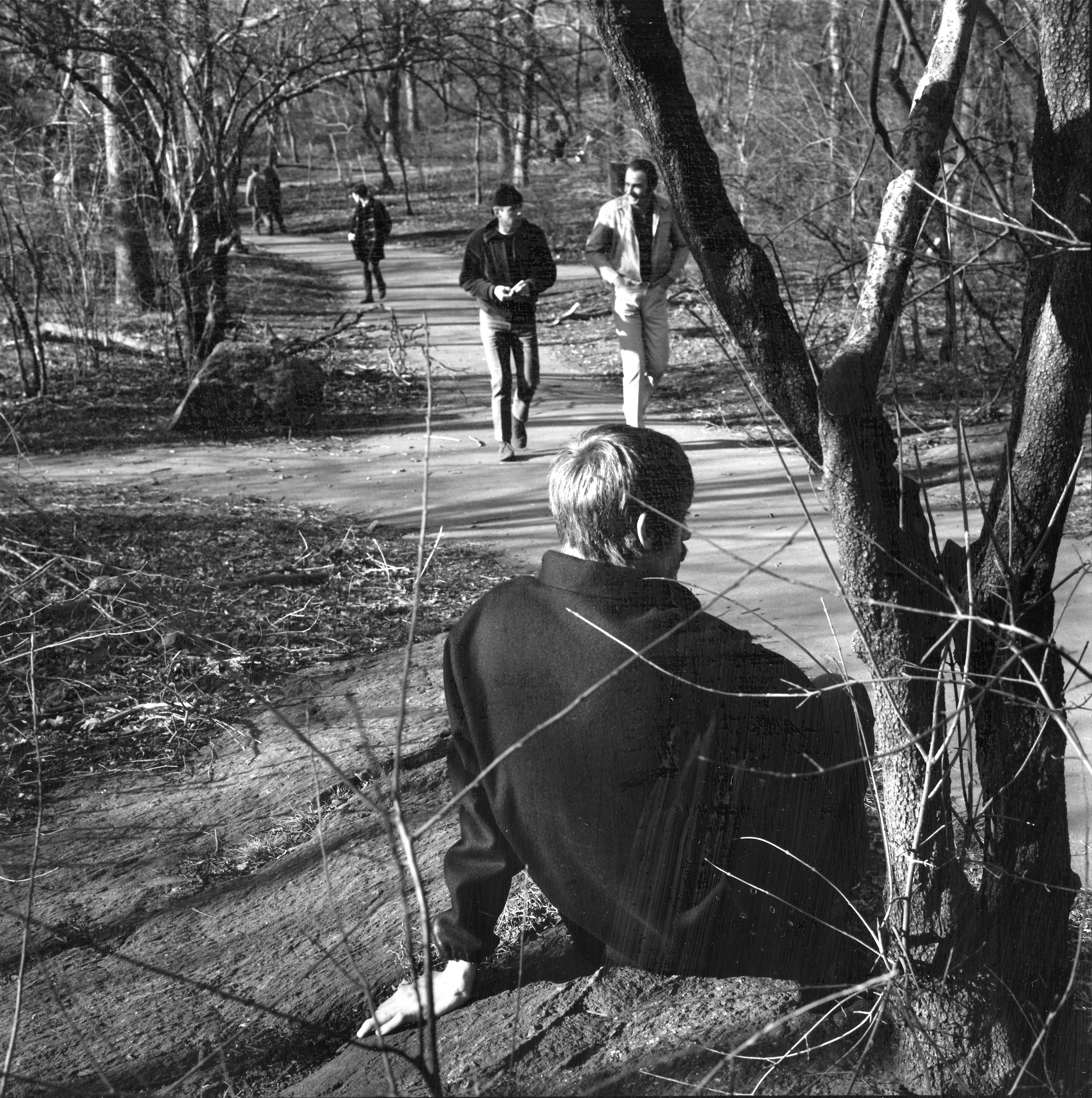 Black and white photograph: man in dark coat sits by bare tree, three people walk on path in background through park setting.