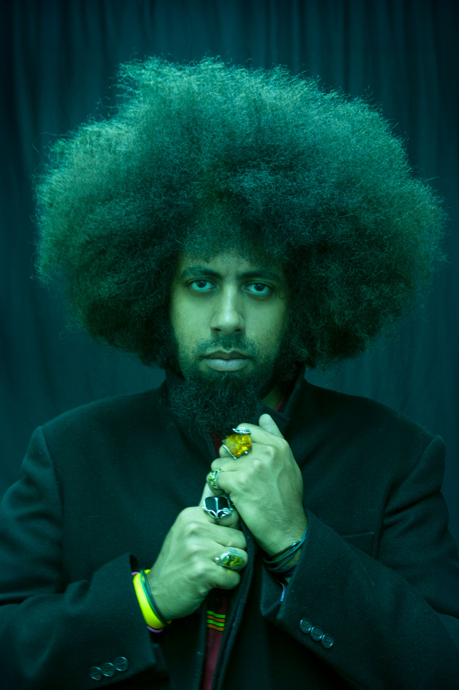Man with large green afro hair and beard wearing black suit, holding microphone, against dark teal background with dramatic lighting.