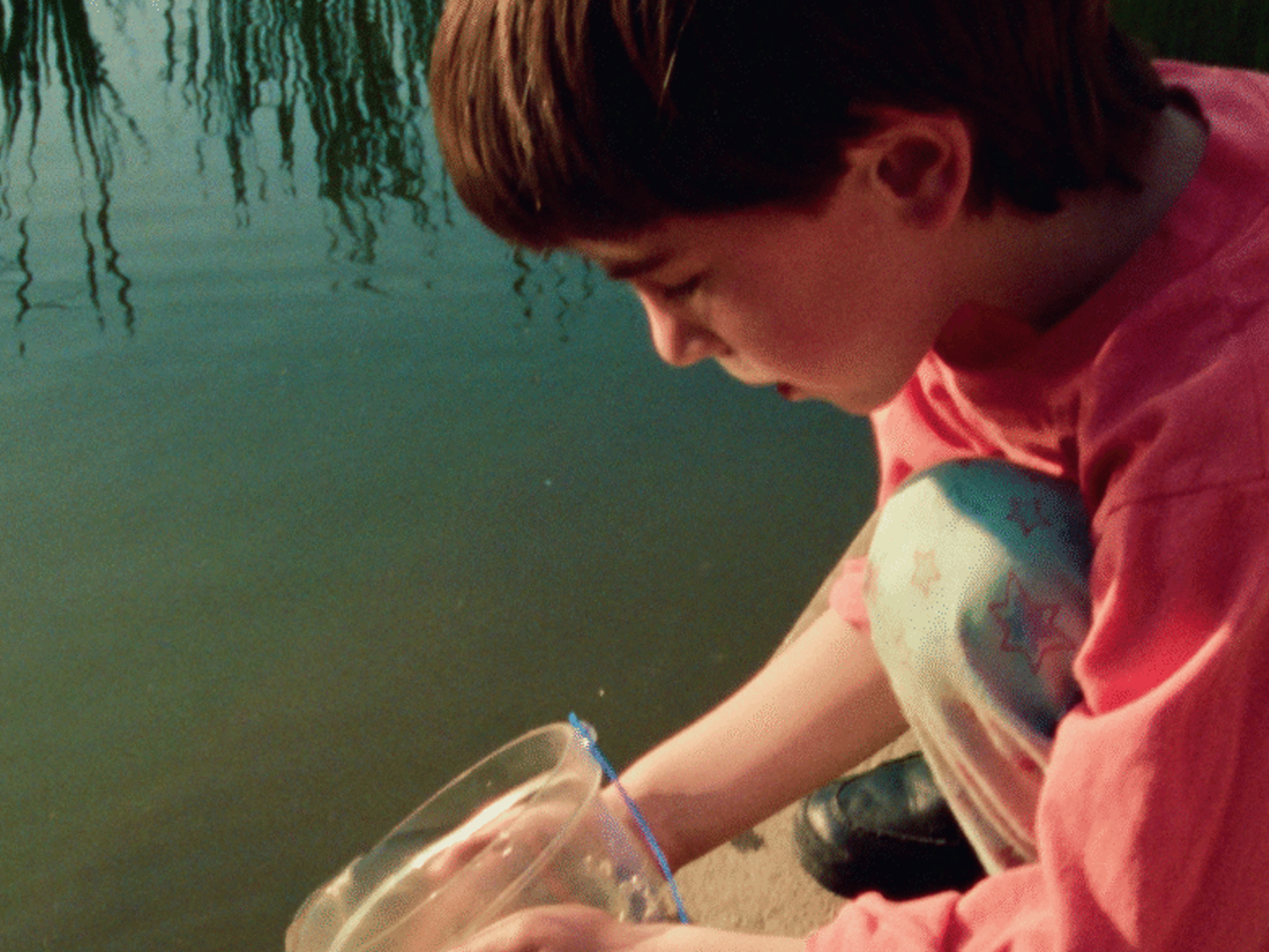 Boy in pink shirt kneeling by water's edge, using clear container to collect water samples.