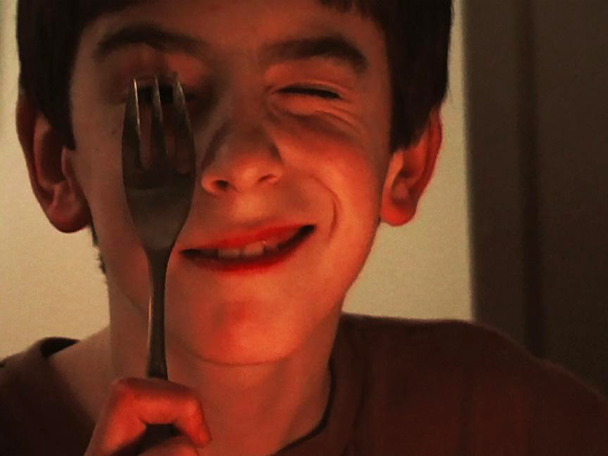 Man holding fork near mouth, smiling broadly. Face illuminated with warm red lighting against dark background.