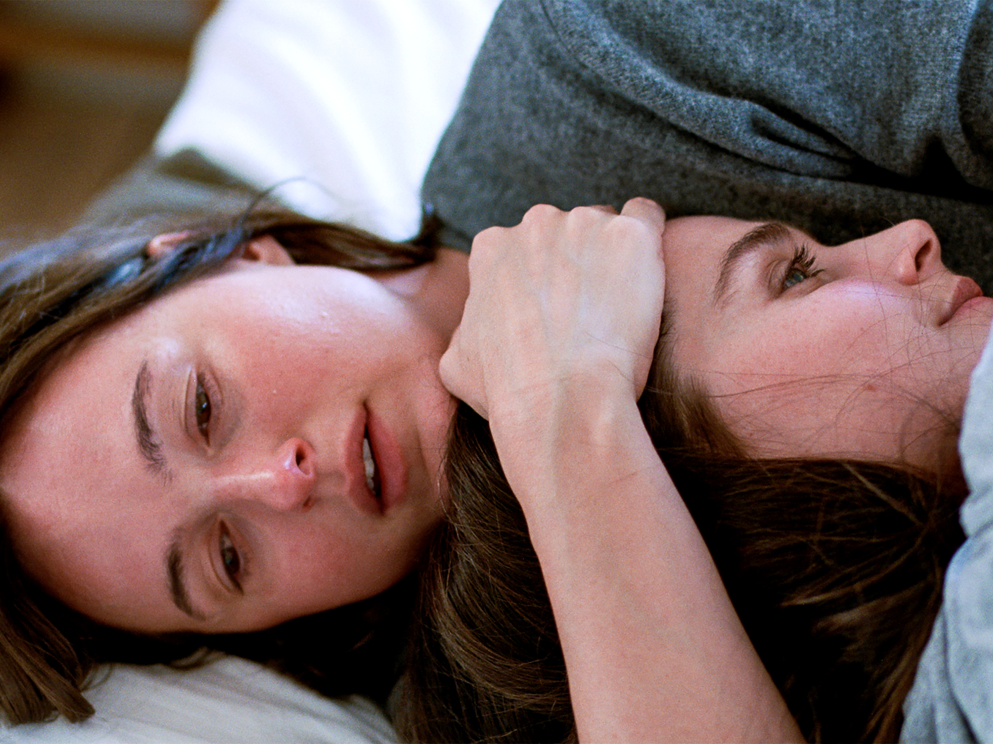 A woman with long brown hair lying on a bed, hugging another woman, her hand on her forehead.