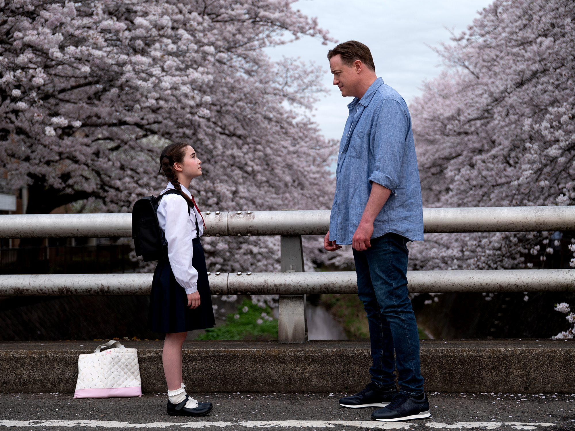 Tall man in blue shirt and young girl in school uniform standing by railing with cherry blossoms in background, white bag on ground.