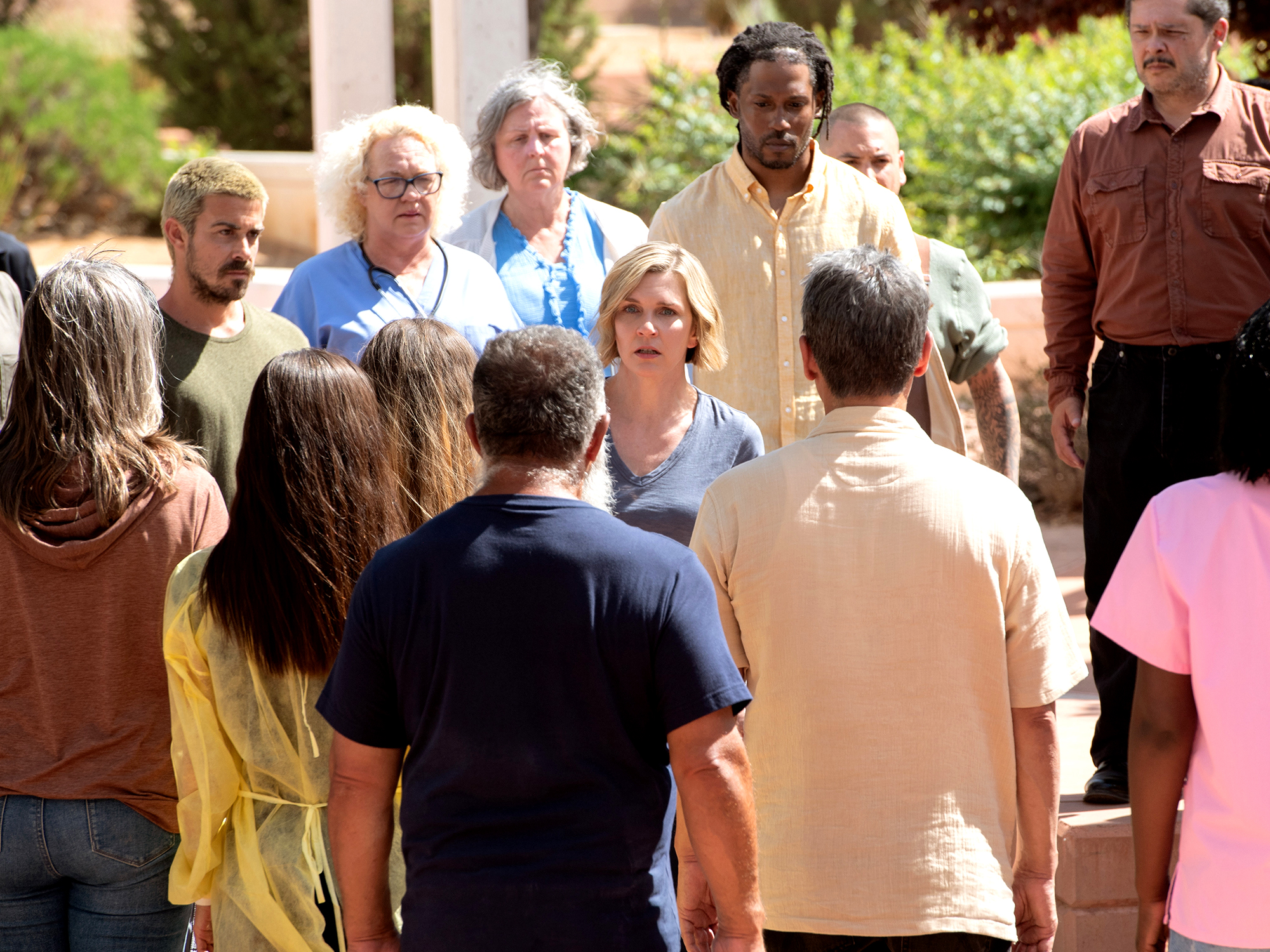 Group of adults gathered outdoors in casual clothing, with blonde woman in centre speaking to others in circle formation.