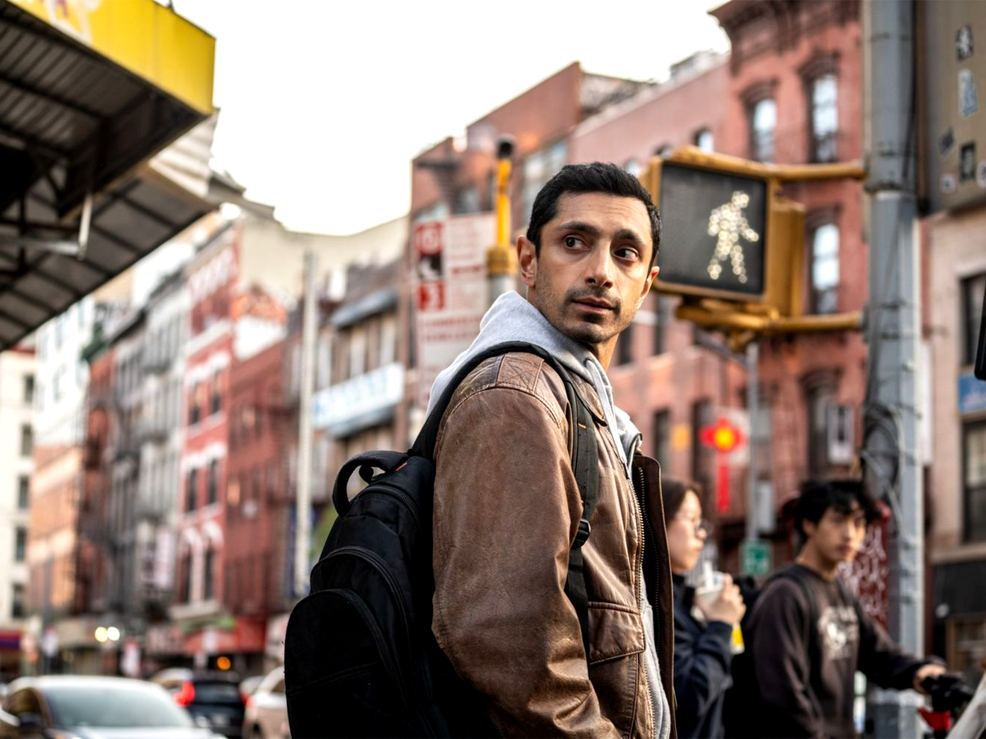Man in brown jacket and rucksack looking over shoulder on busy street with colourful brick buildings and traffic lights.