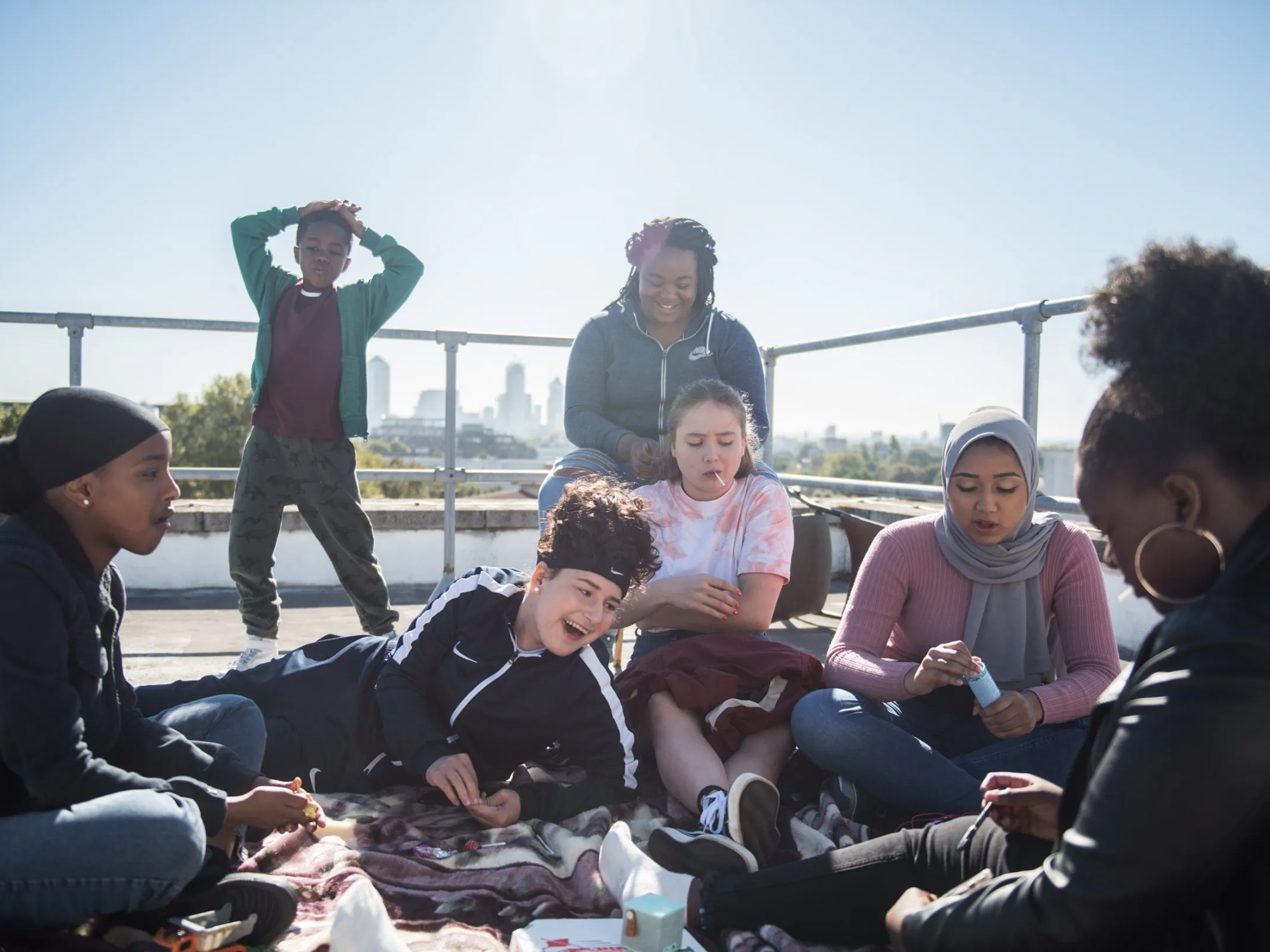 A group of teenage girls sit on a roof in the sunshine, laughing and smiling.