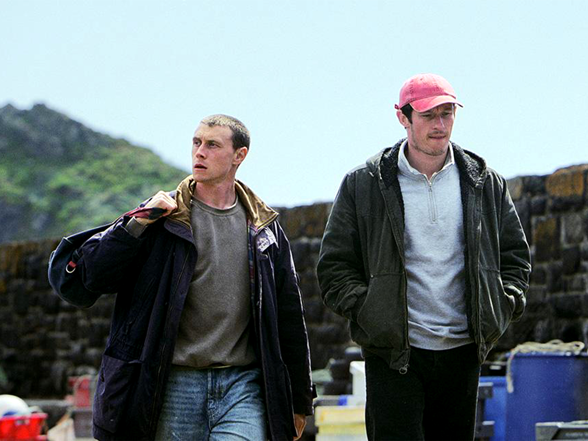 Two men walking outdoors, one in dark leather jacket carrying clothing, other in green jacket and pink cap, mountains in background.