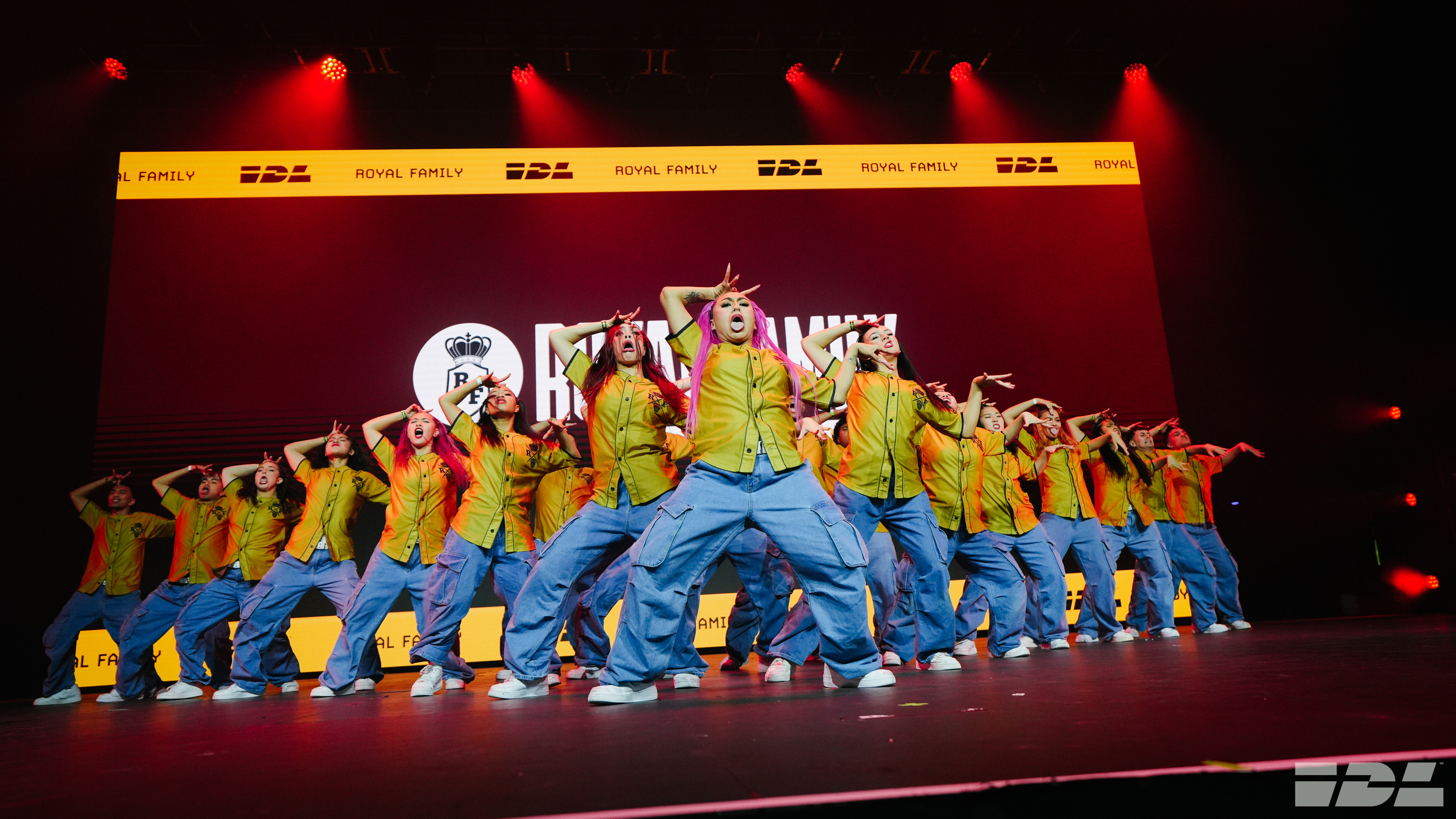 Group of performers in yellow tops and blue bottoms on stage with red lighting and yellow backdrop displaying sponsor logos.
