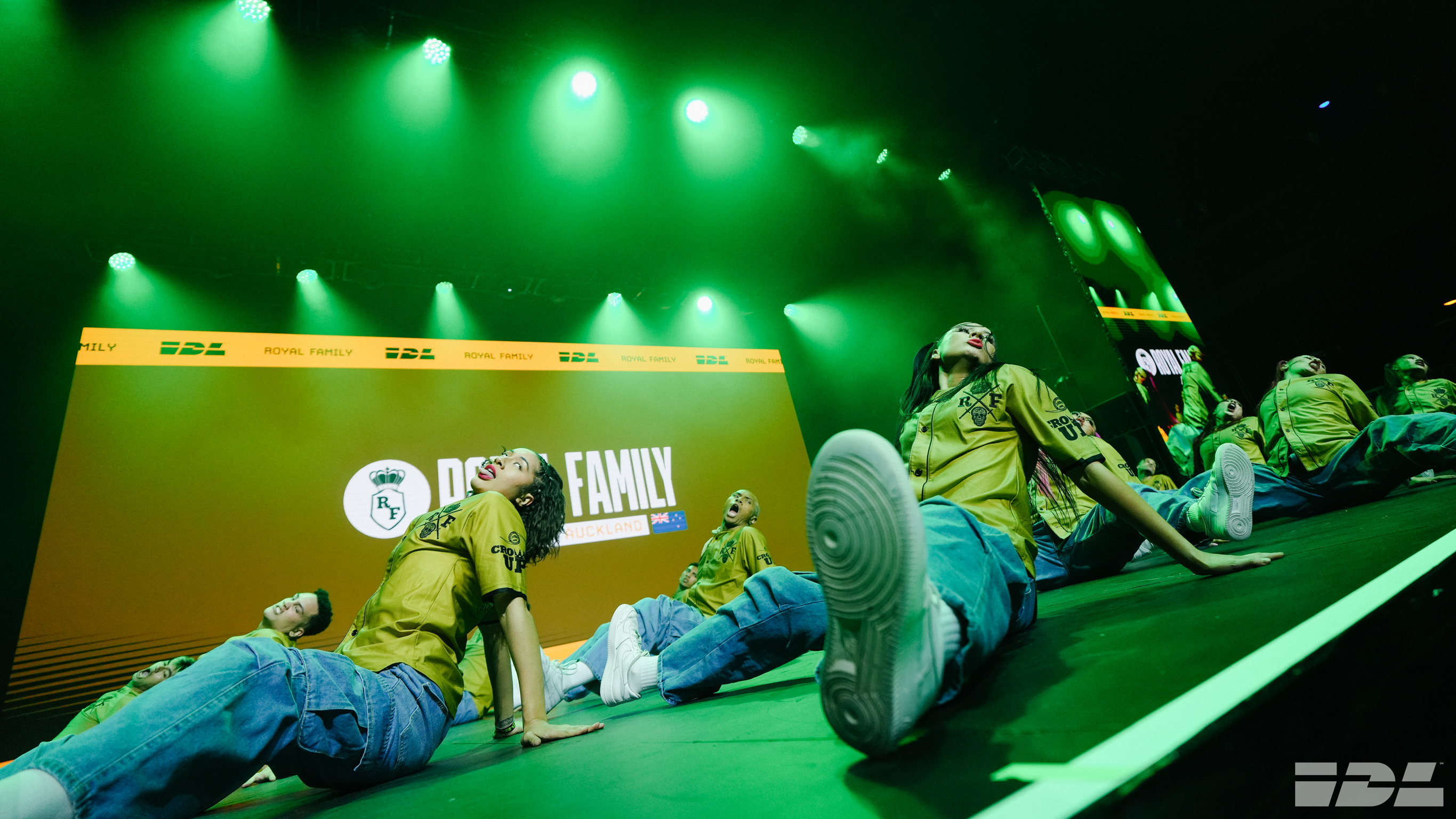 Stage lit in bright green light with performers in casual clothing lying and sitting on floor. Yellow banner with "FAMILY" text visible.