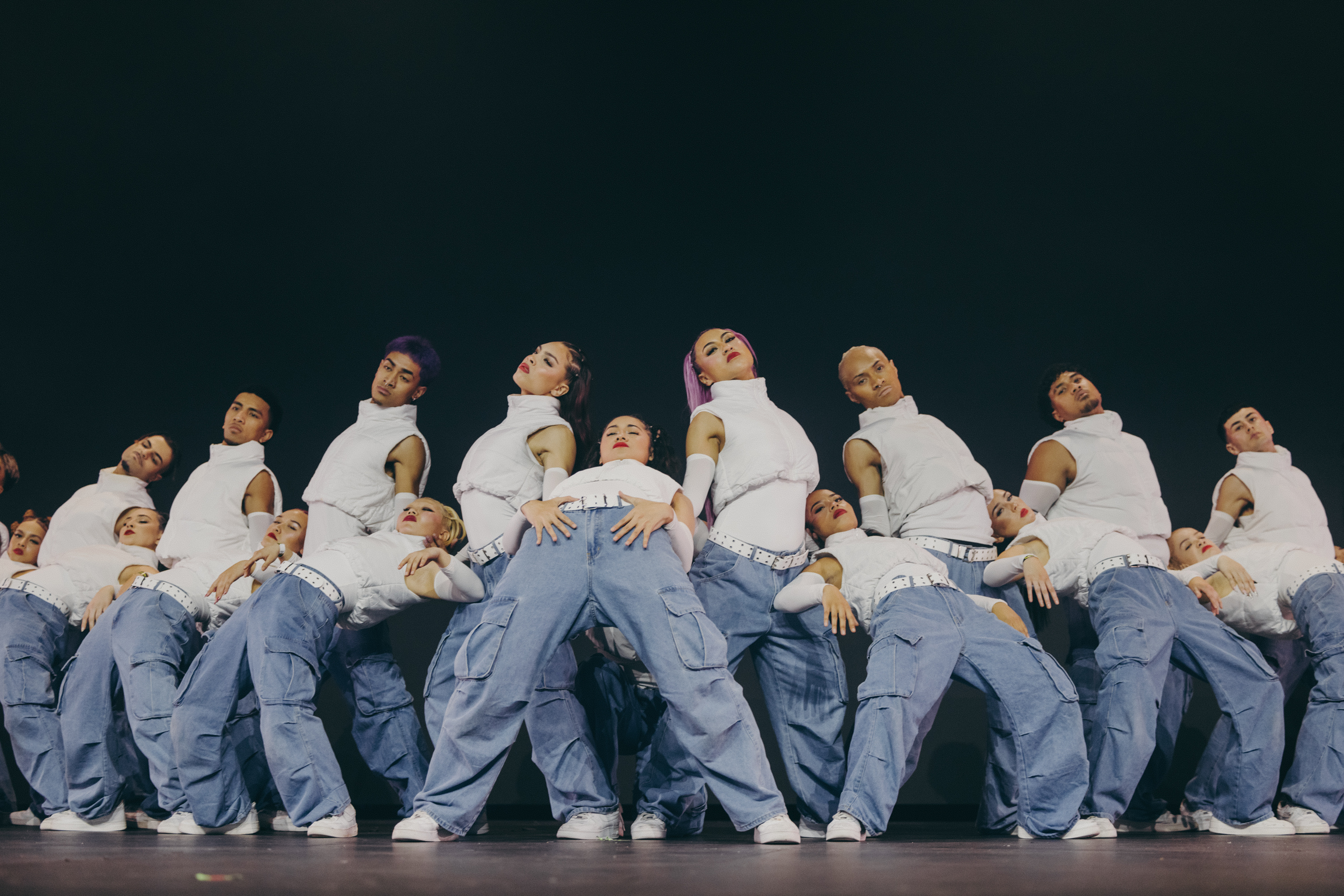 Group of performers in white tops and blue trousers arranged in formation on stage against dark background.