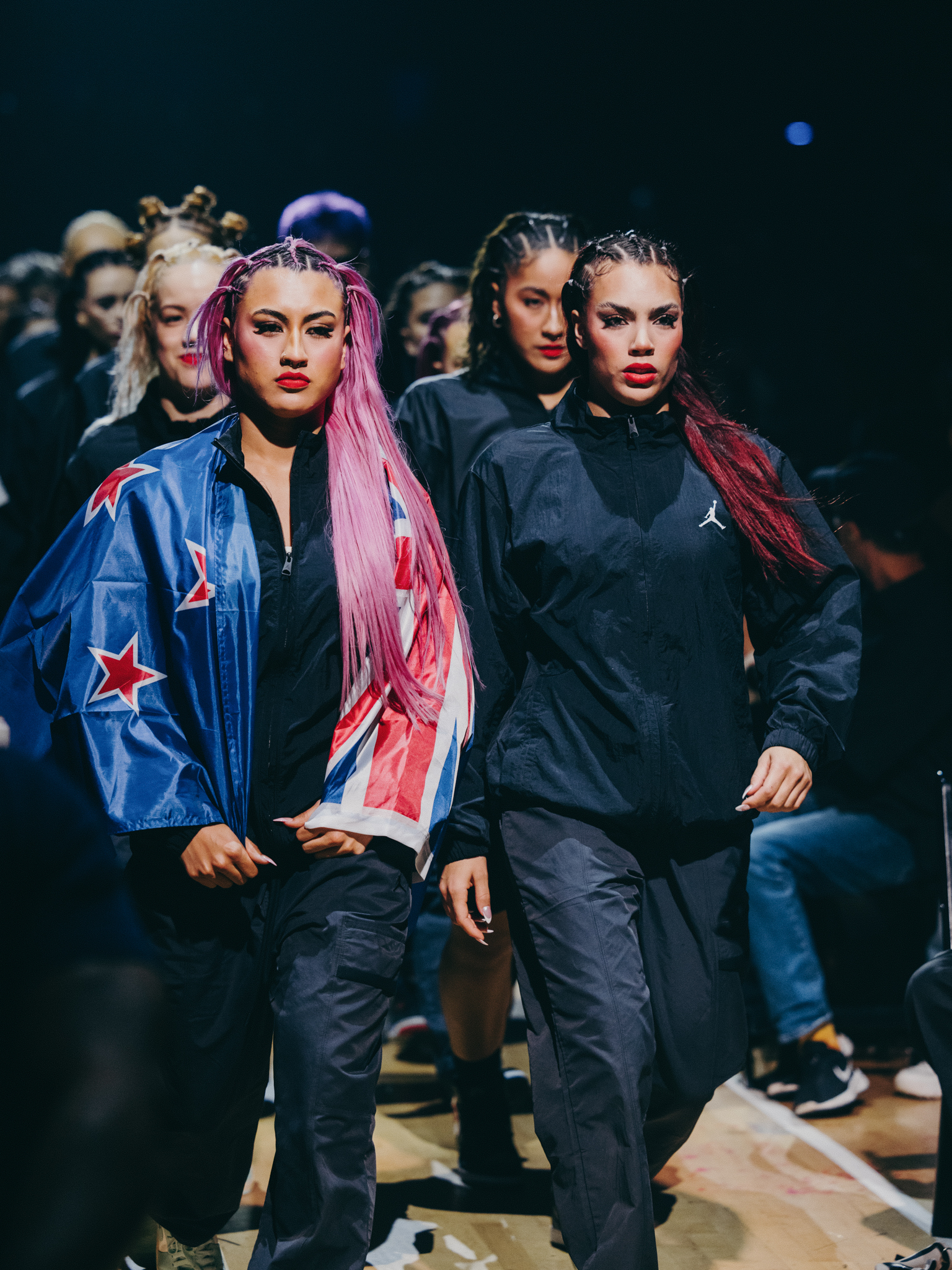 Two performers with pink and red braided hair wearing blue star-patterned and black tracksuits, surrounded by crowd in dark setting.