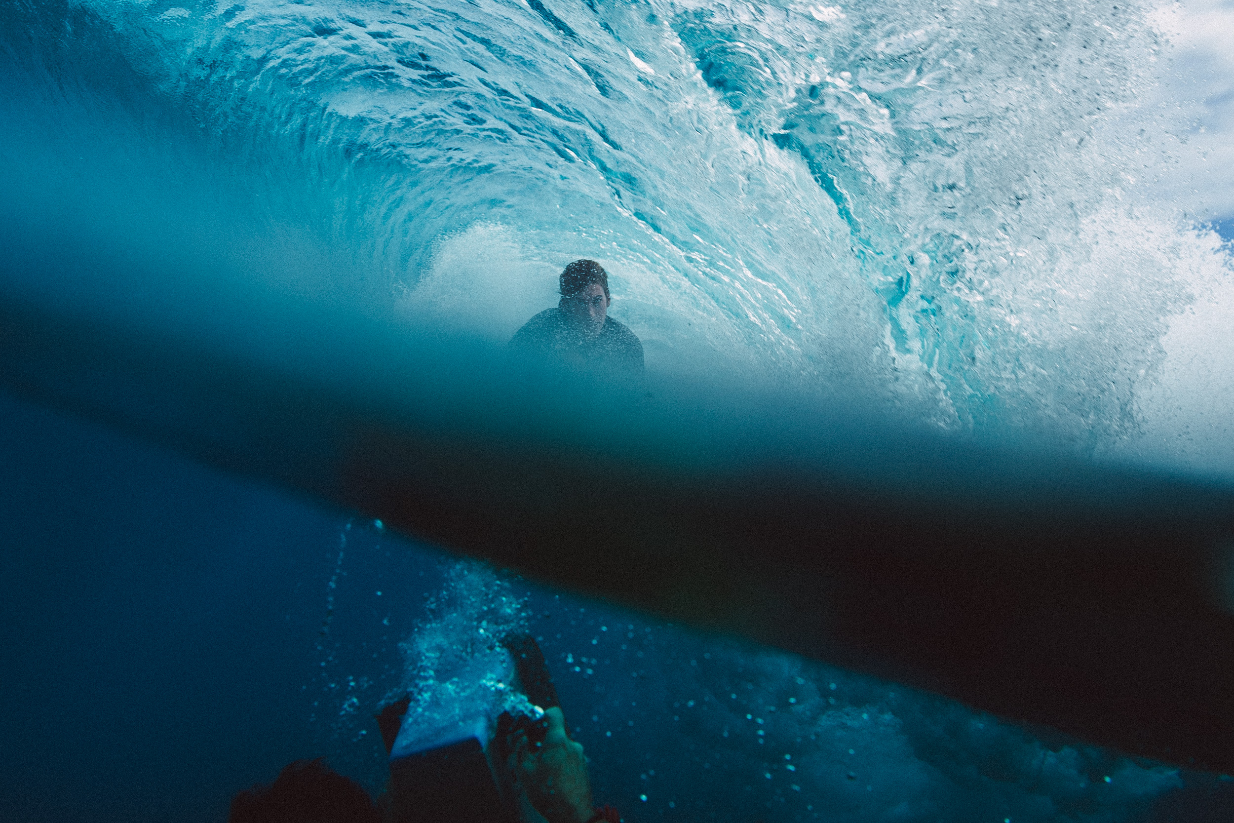 Underwater view of person swimming beneath a breaking wave, with turquoise water, white foam, and dark silhouettes against bright surface.
