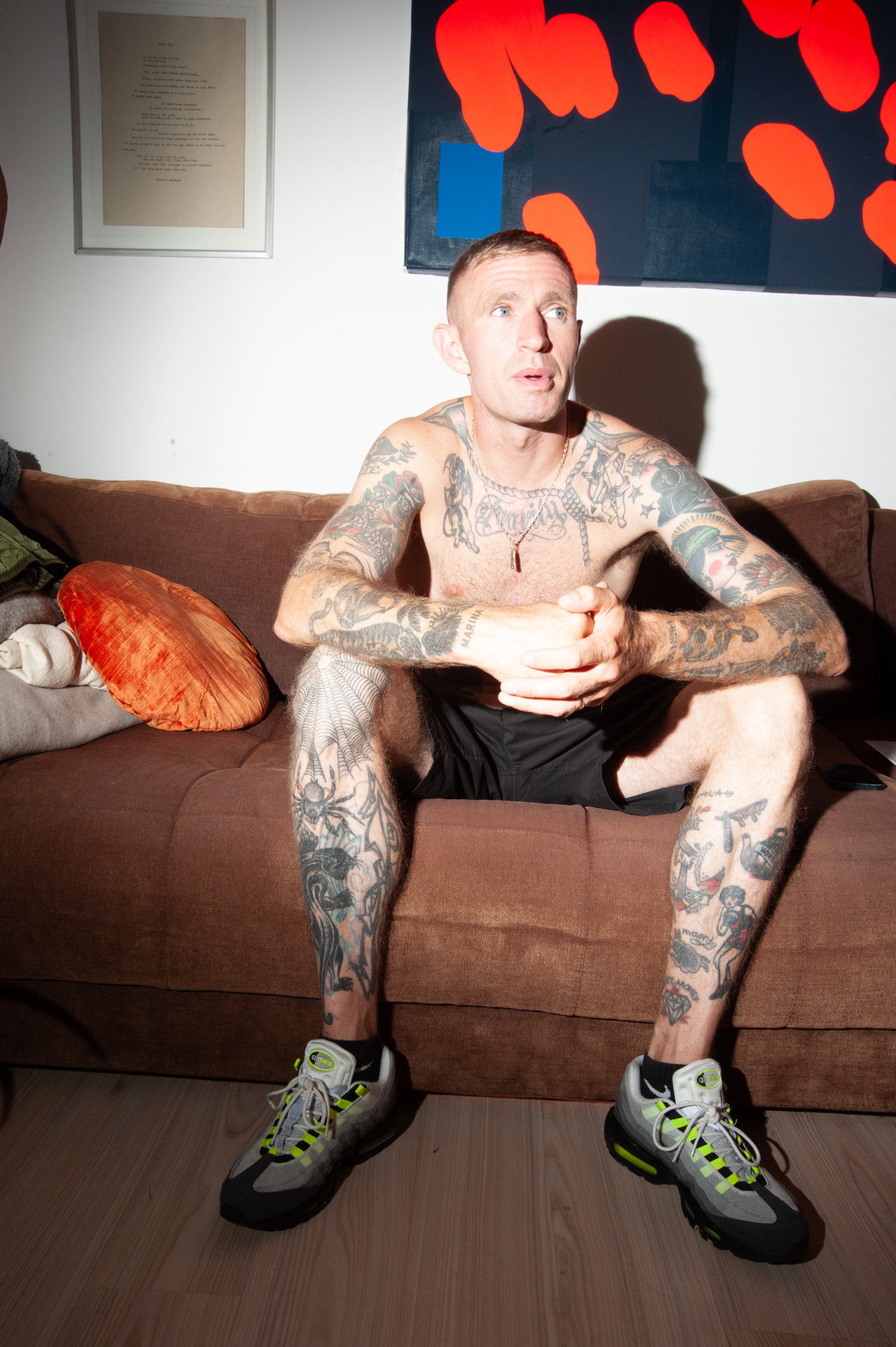 Heavily tattooed man in shorts and trainers sits on brown sofa, holding mobile phone. Abstract painting with red shapes on dark background hangs above.