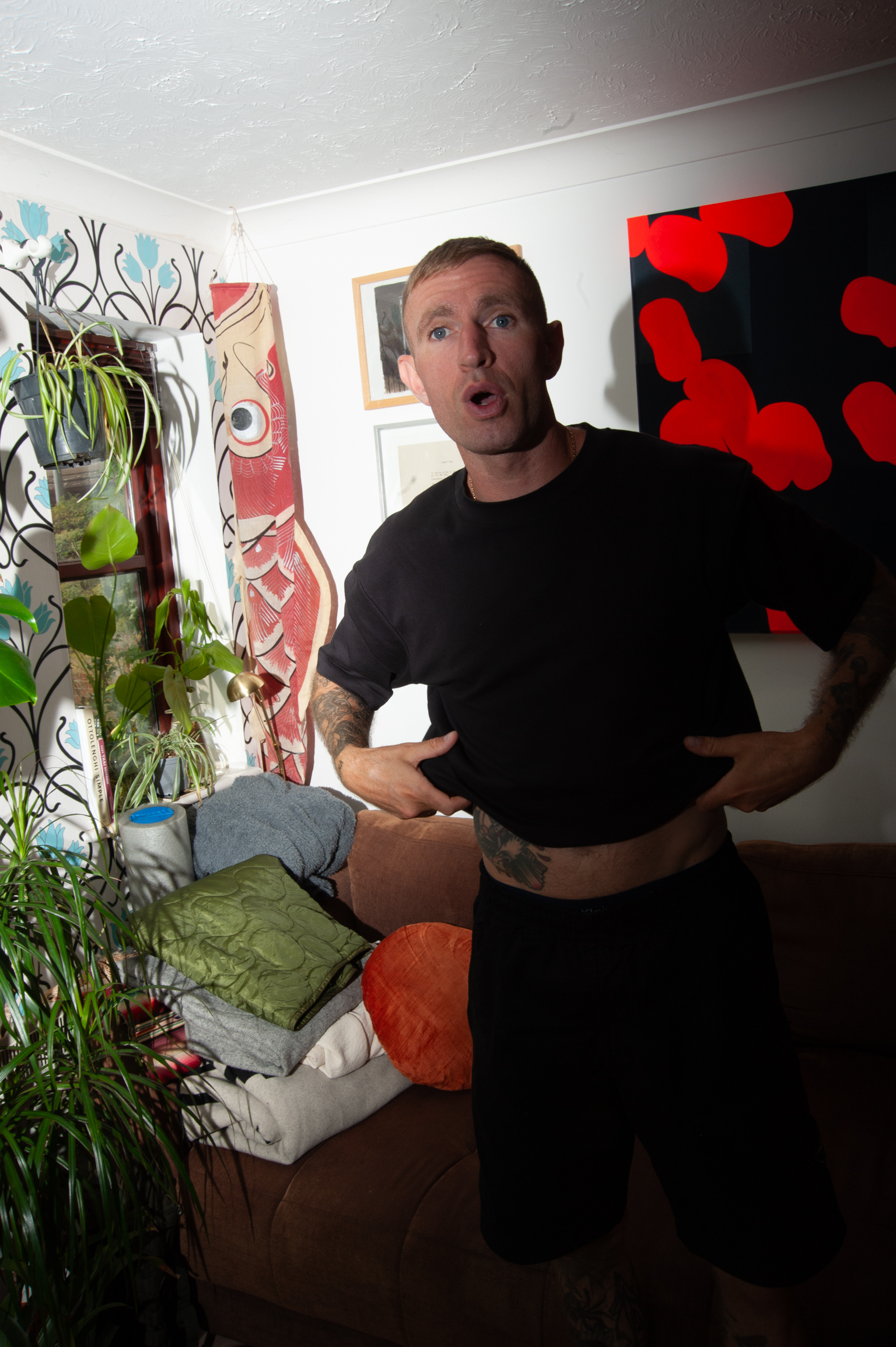 Man in black t-shirt gesturing with surprised expression in room with colourful artwork on walls and green plants.