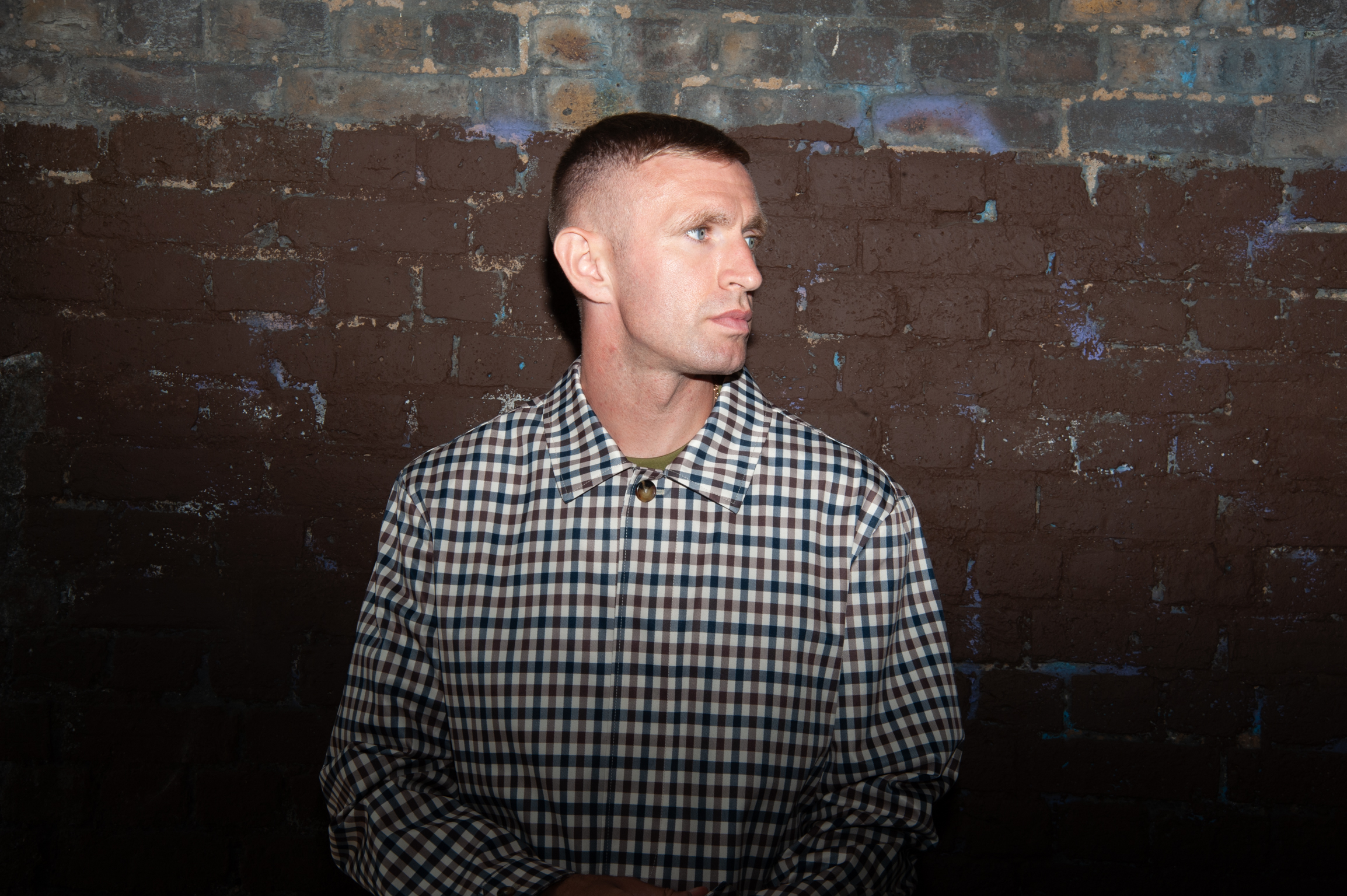 Man in black and white checked shirt looking to the right against weathered brick wall with peeling paint.