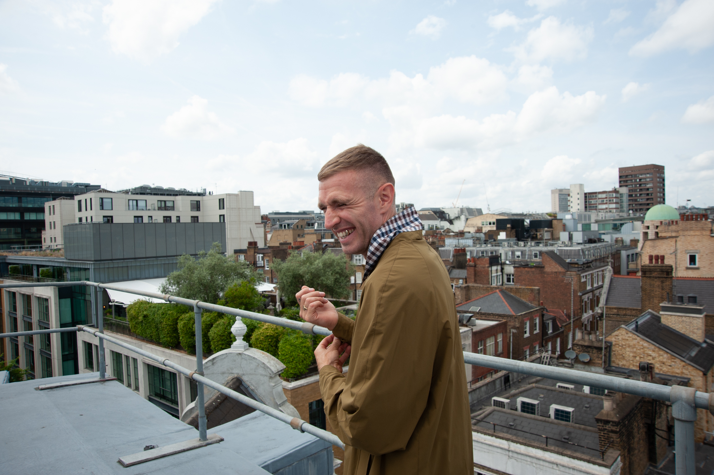 Man in brown coat and striped scarf standing on rooftop terrace overlooking urban cityscape with mixed residential and commercial buildings.