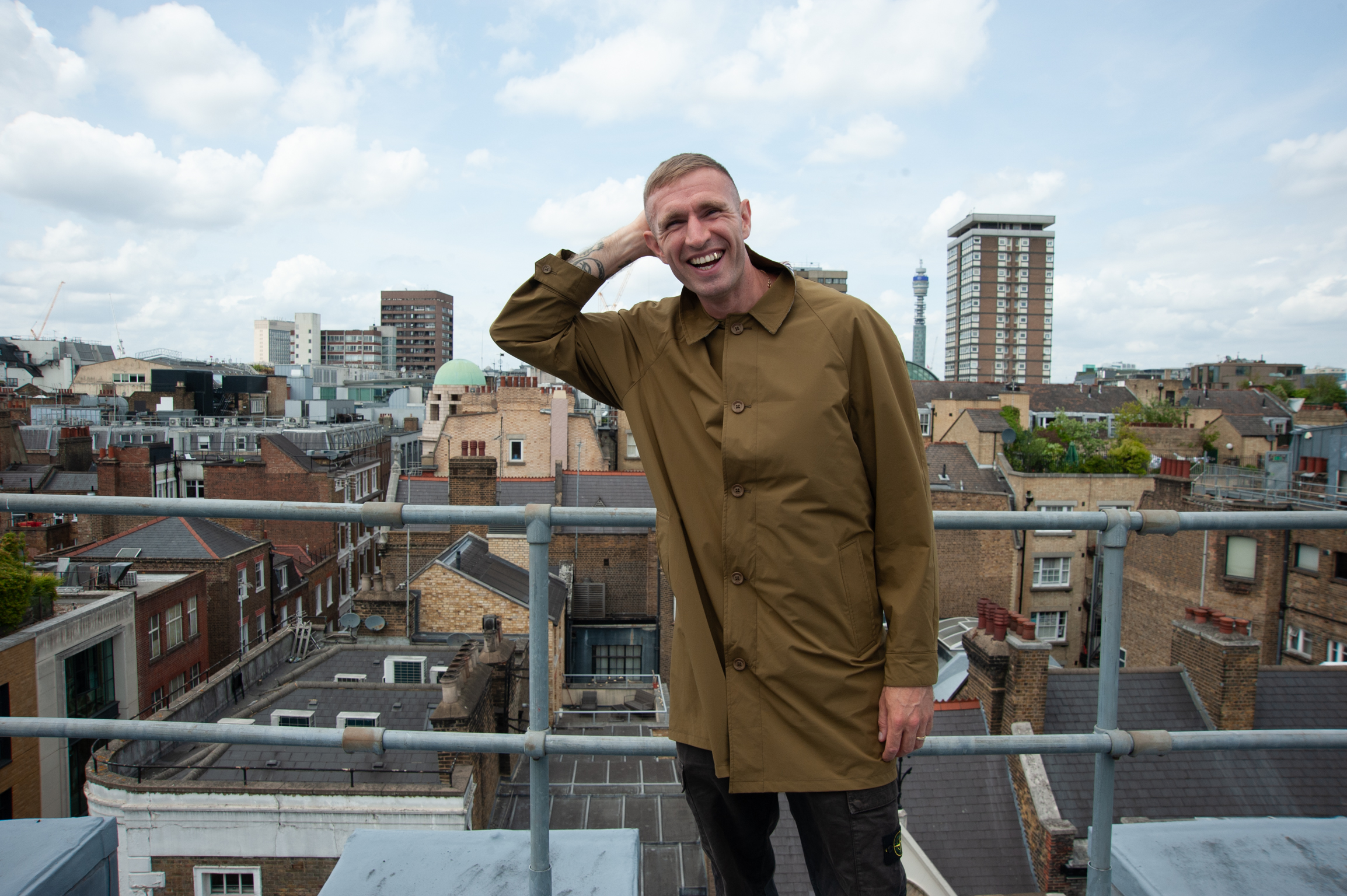 Man in khaki coat saluting on rooftop with urban cityscape of mixed residential and commercial buildings in background.