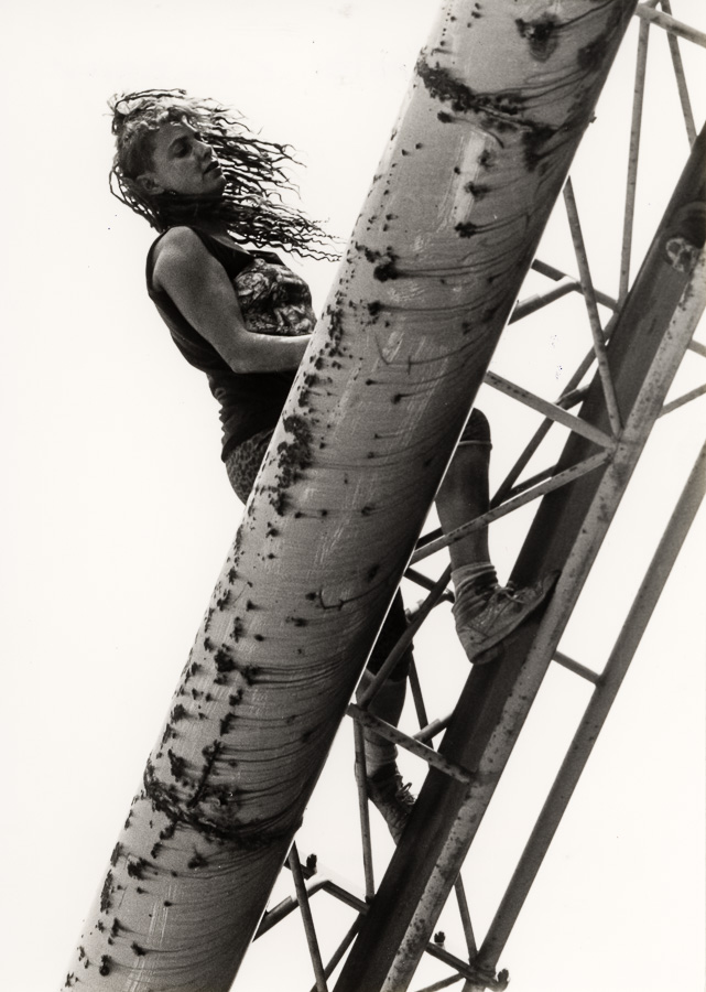 Woman with long dark hair climbing industrial metal tower structure, viewed from below against pale sky in monochrome.