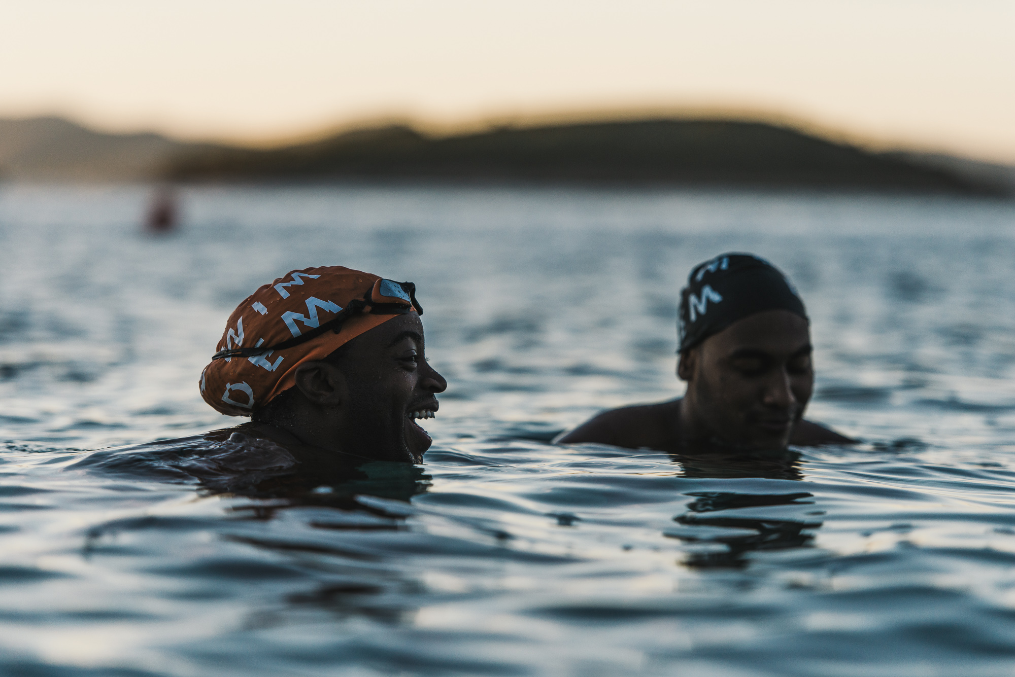 Two people swimming in calm water, one wearing a patterned headscarf, with distant hills and pale sky in background.
