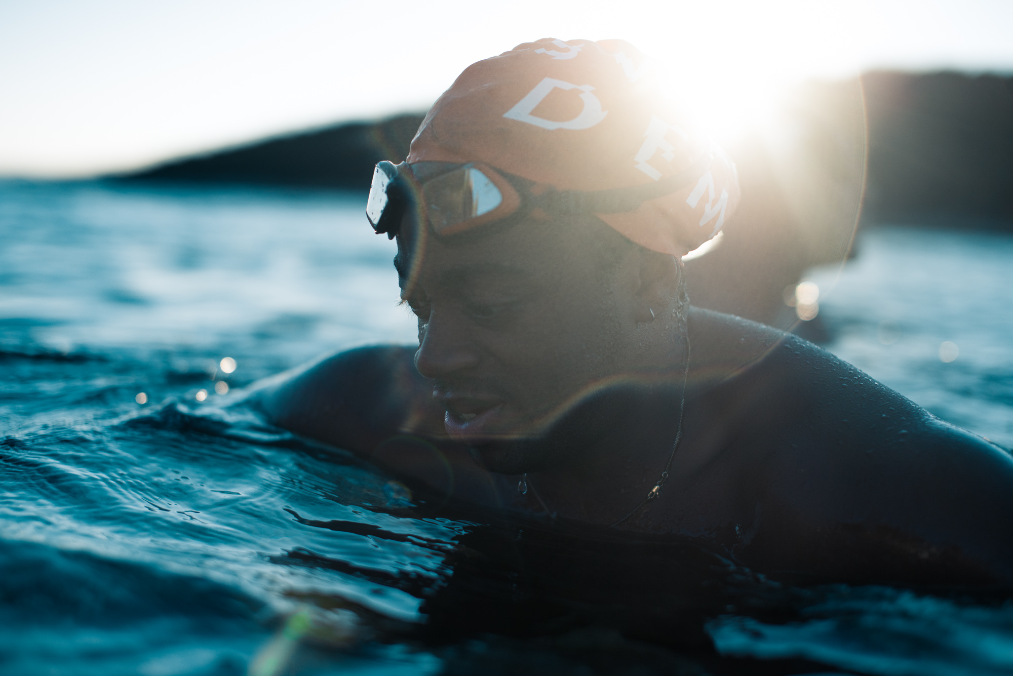 Person in swimming goggles and cap in blue water with bright sunlight creating lens flare, dark hills visible in background.