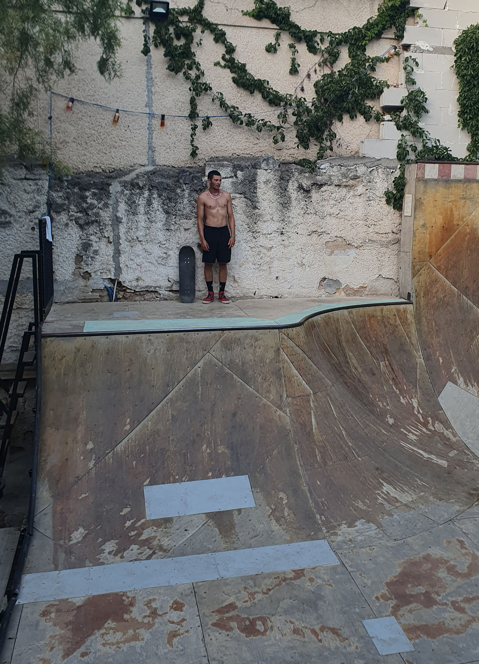 Shirtless man stands beside empty concrete skate bowl with weathered walls, green ivy hanging overhead, and metal railings.