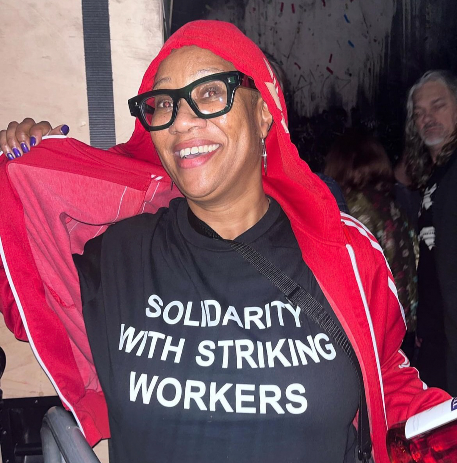 Person wearing black glasses and red hoodie holds red jacket. Black t-shirt reads "SOLIDARITY WITH STRIKING WORKERS" in white text.