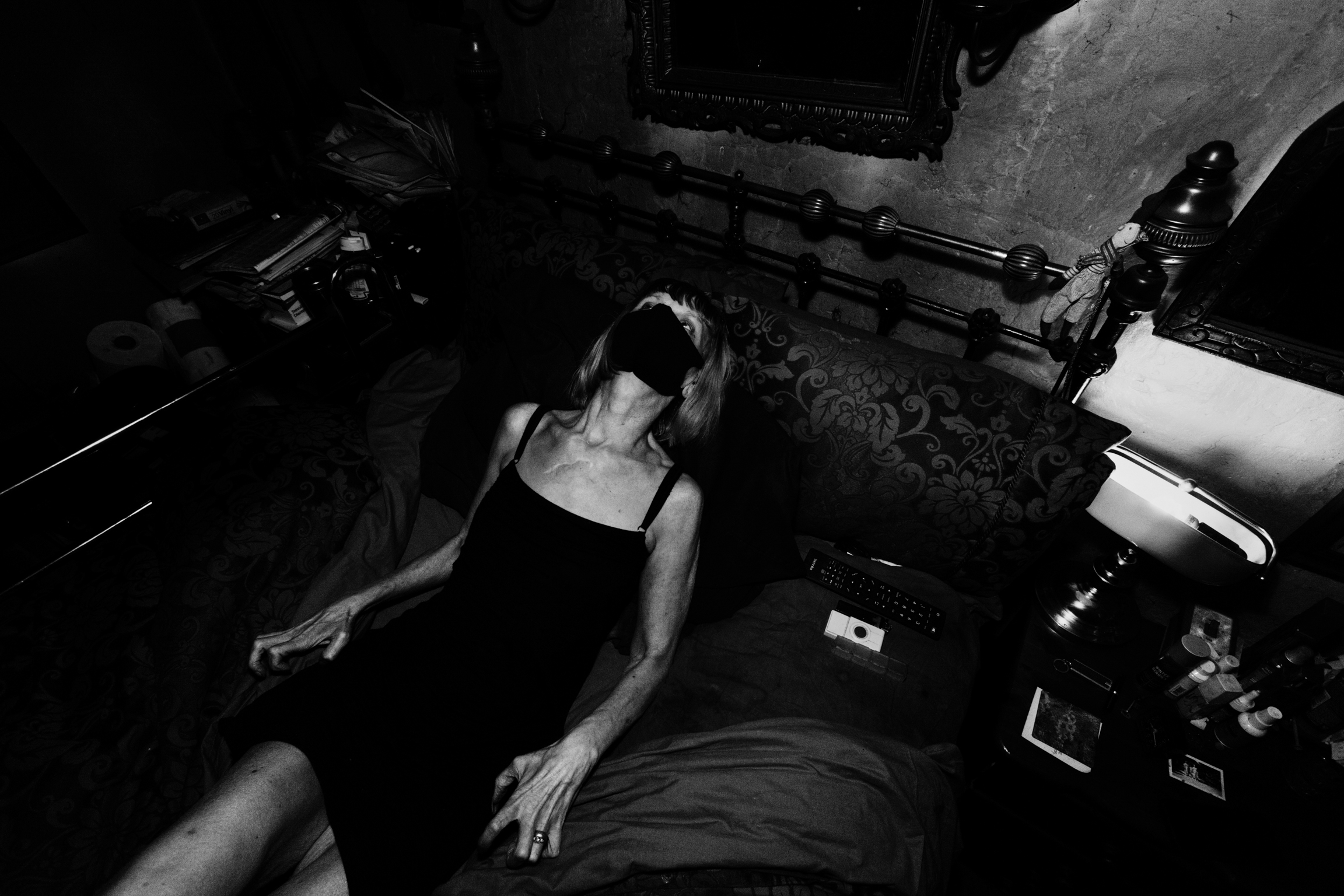 Black and white image showing person in dark clothing lying on floor amongst scattered papers and objects in cluttered room.