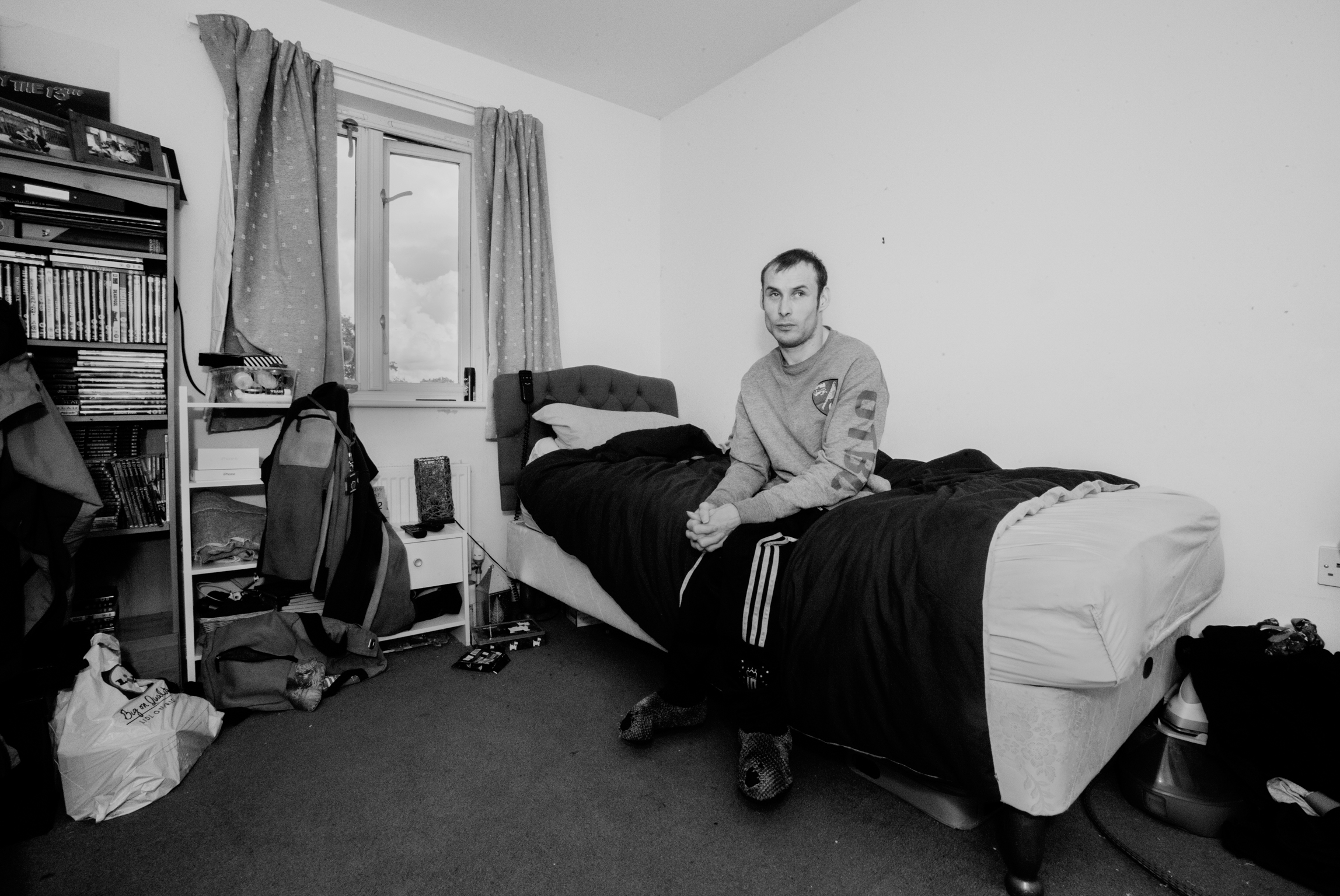 Man sitting on bed in sparse bedroom with entertainment system, curtained window, and scattered belongings on floor.