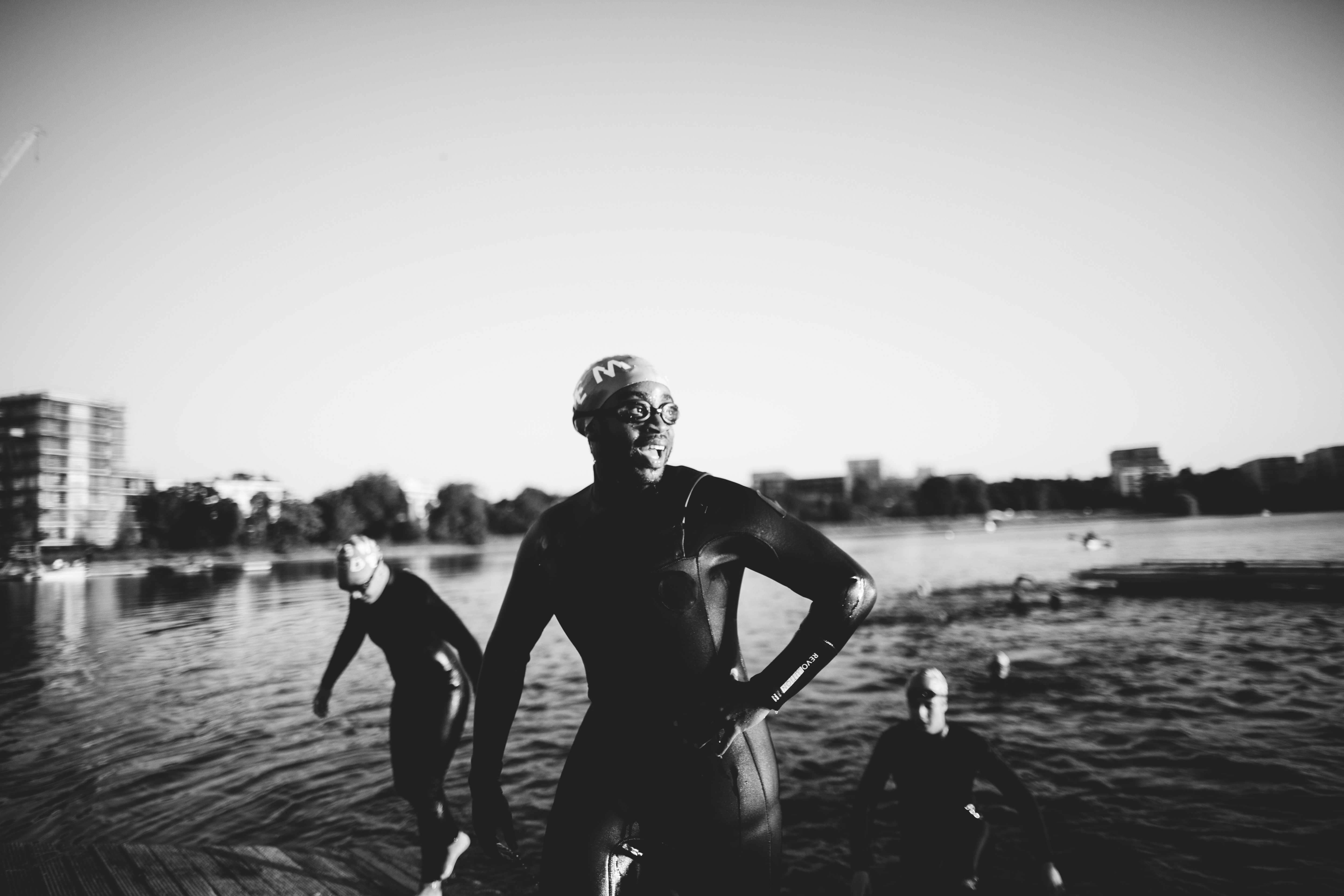 Person in wetsuit holding swimming gear stands by water's edge with buildings visible across lake in black and white image.