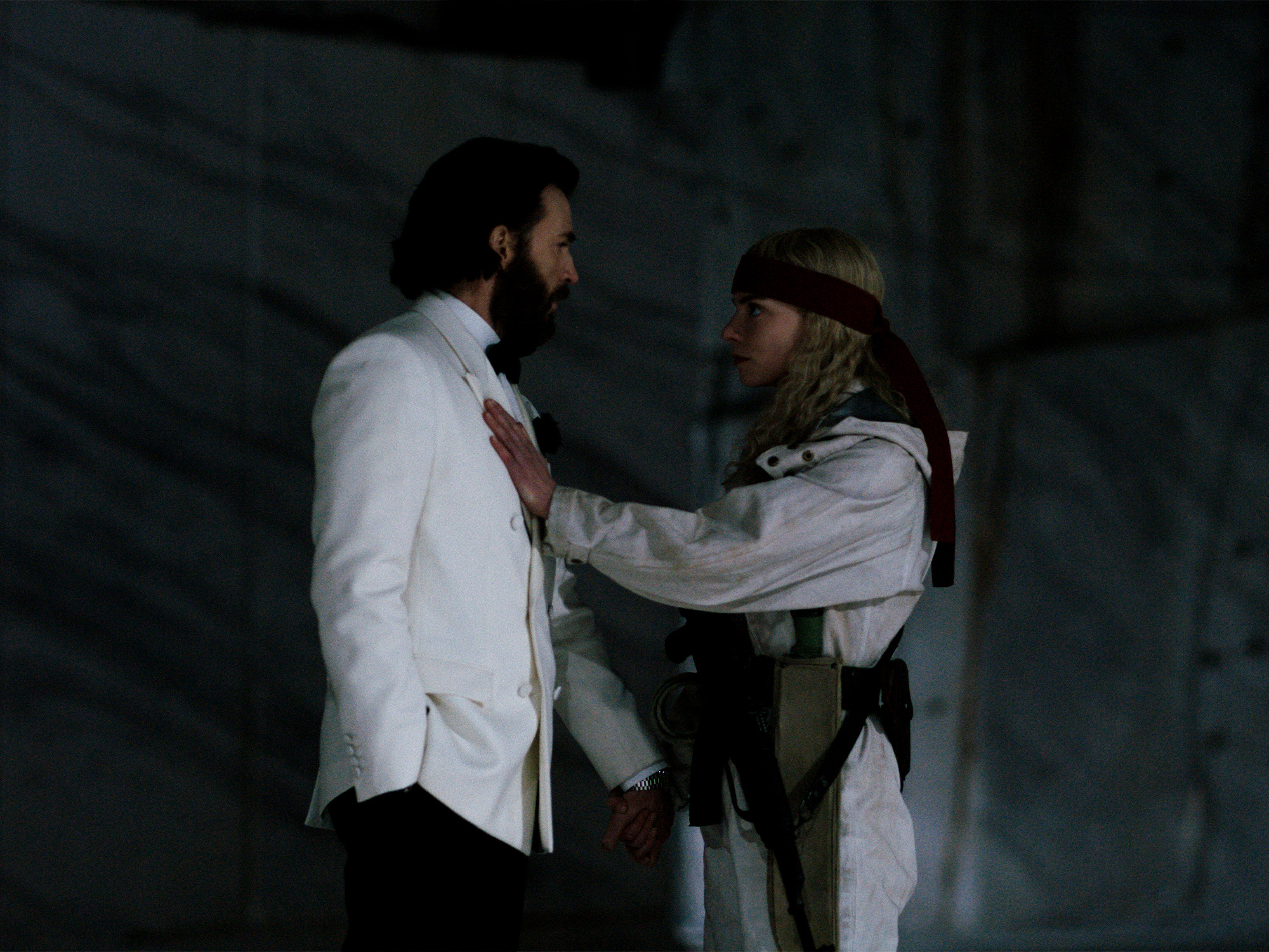 Two men in white shirts on dark stage, one wearing headband, hands positioned on each other's shoulders in dim lighting.
