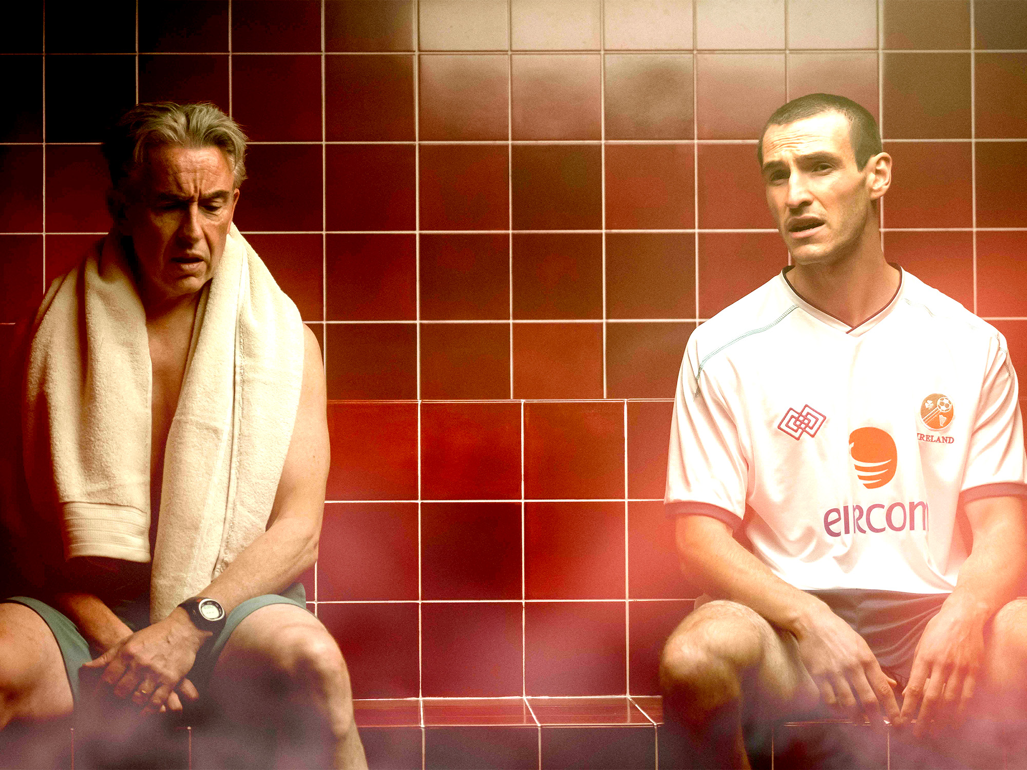 Two men sitting on bench in changing room with red tiled wall. Left man wears towel, right man in white football shirt.