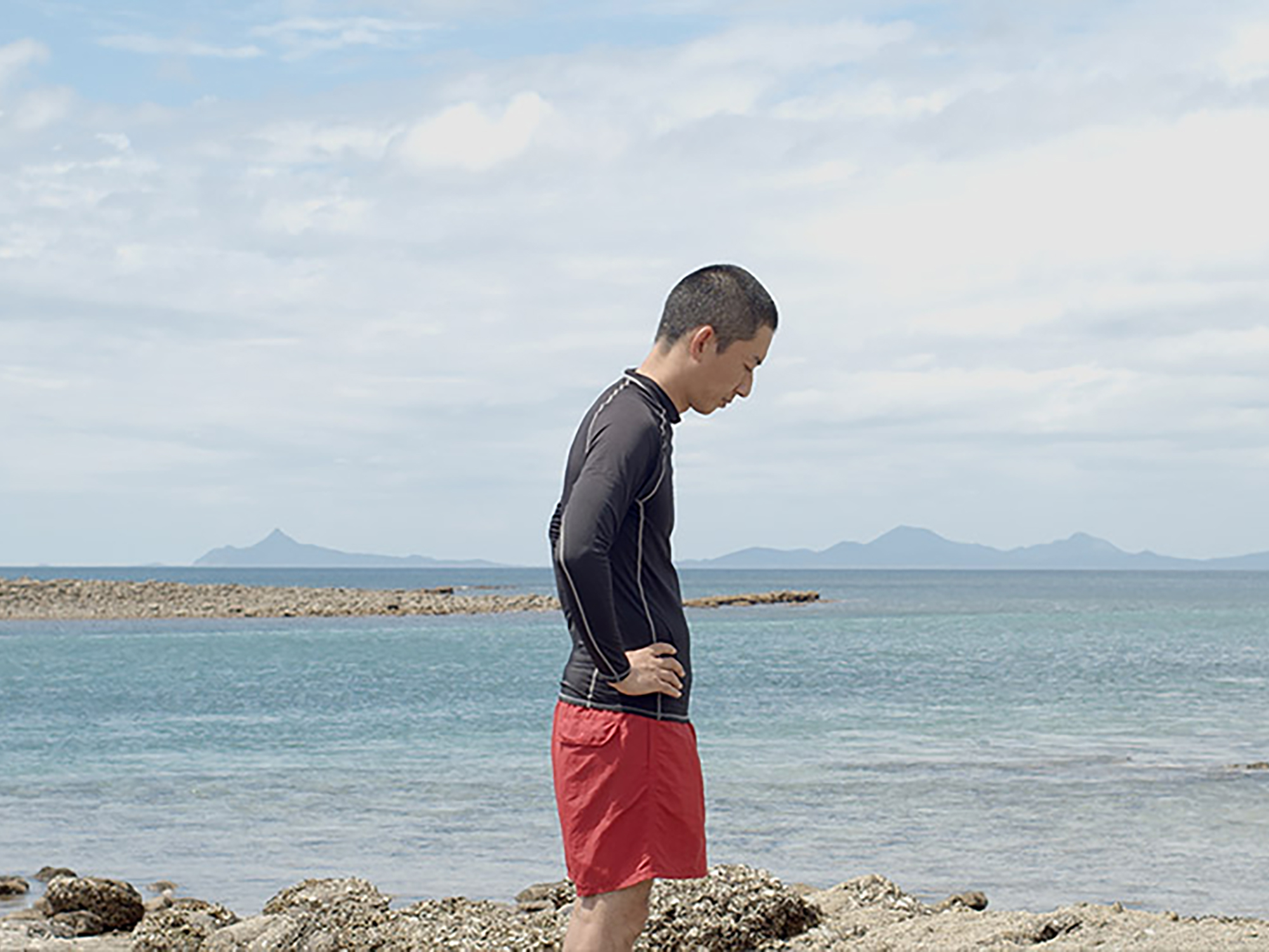 Man in black long-sleeved shirt and red shorts standing on rocky beach, looking down. Blue sea and cloudy sky behind him.